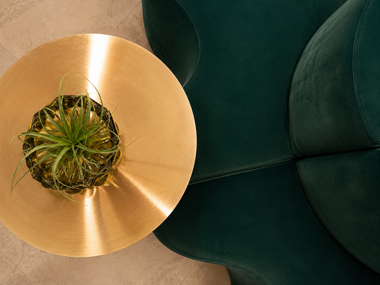 Top-down view of a round gold-colored side table with a glass bowl containing a green succulent plant, next to a dark green upholstered armchair with rounded armrest.