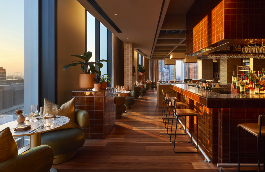 Interior of a modern upscale restaurant or bar with large windows, a curved bar counter with bottles, and seating area with tables and chairs, illuminated by warm lighting at sunset.