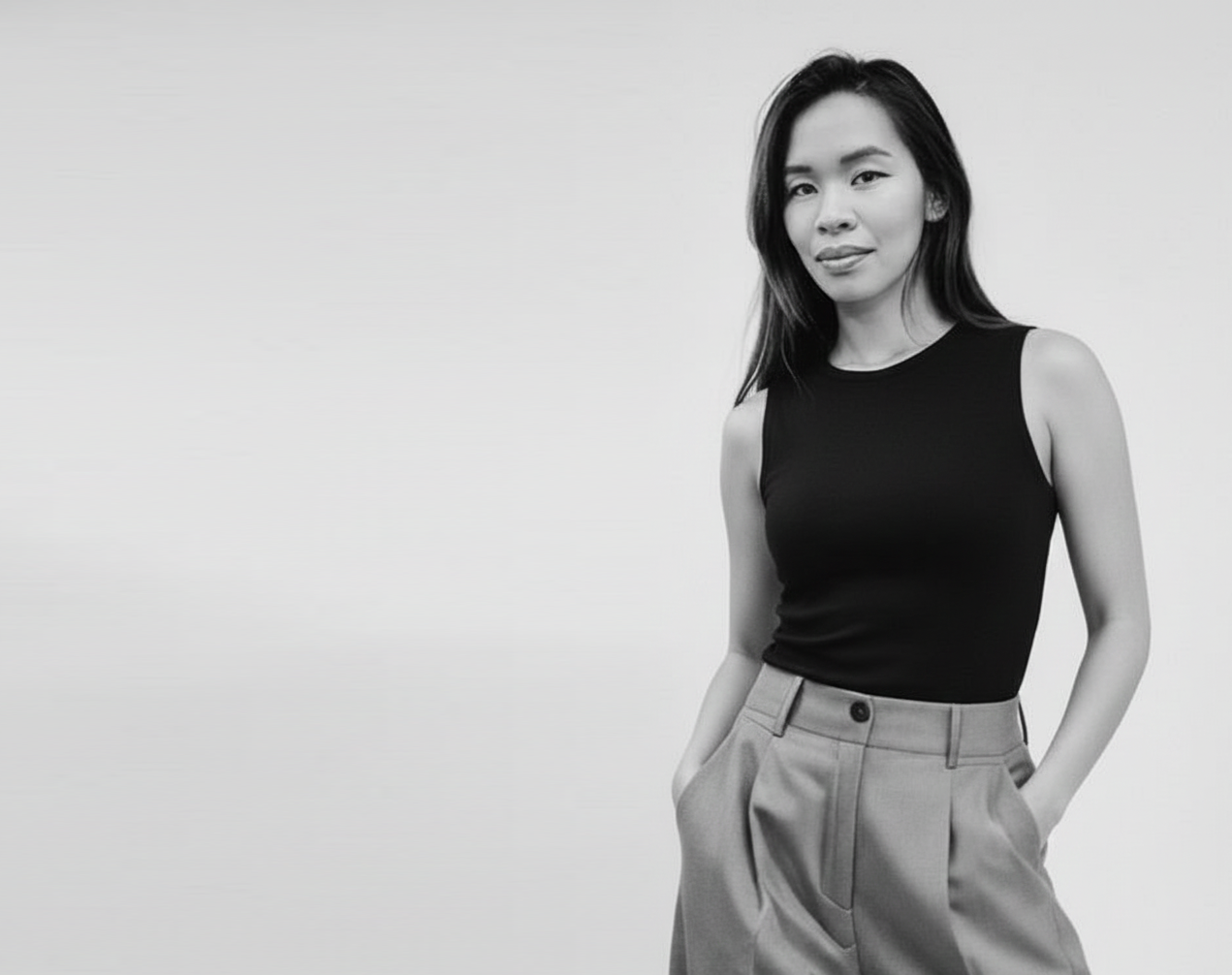Black and white photograph of a woman standing with her hands in the pockets of her high-waisted trousers, wearing a sleeveless black top, standing against a plain light-colored background.