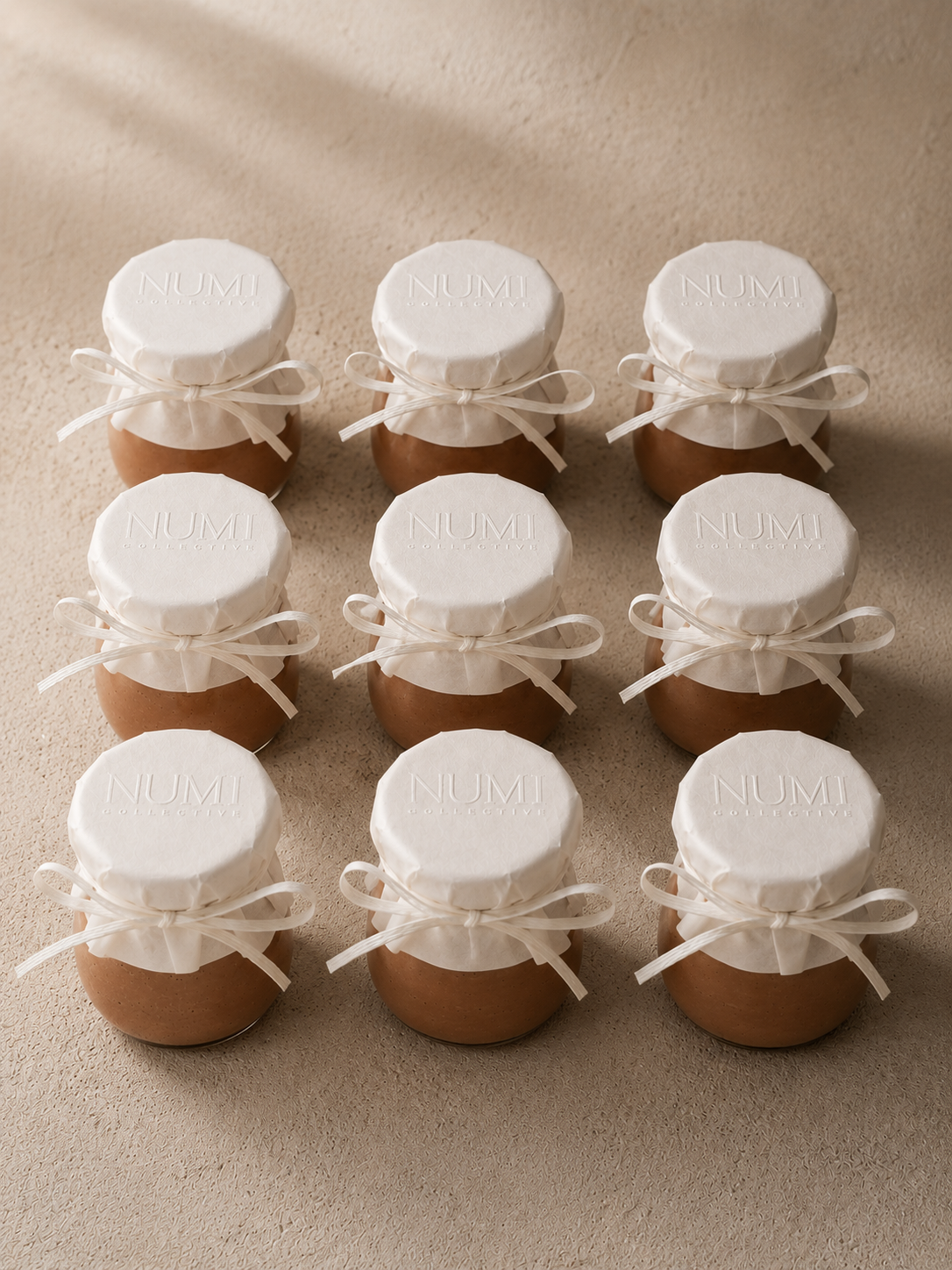Nine brown eggs arranged in three rows on a beige surface, each egg topped with white paper covers with the words 'NUMI COLLECTIVE' embossed on them, tied with white ribbons.