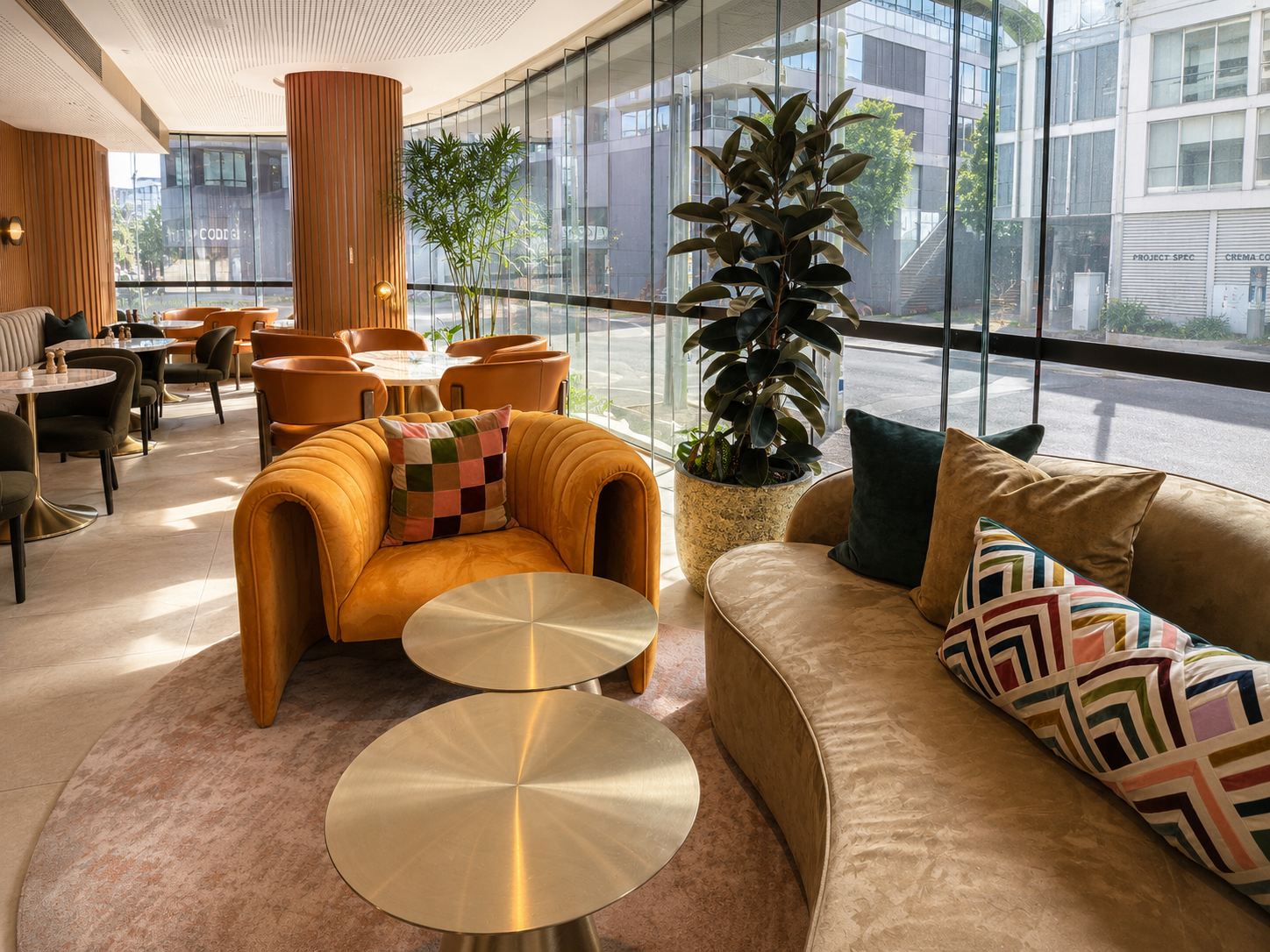 Modern hotel or cafe lounge with large windows, leather and fabric armchairs, a sofa, and several round metallic tables, decorated with colorful pillows, large potted plants, and a city street visible outside.