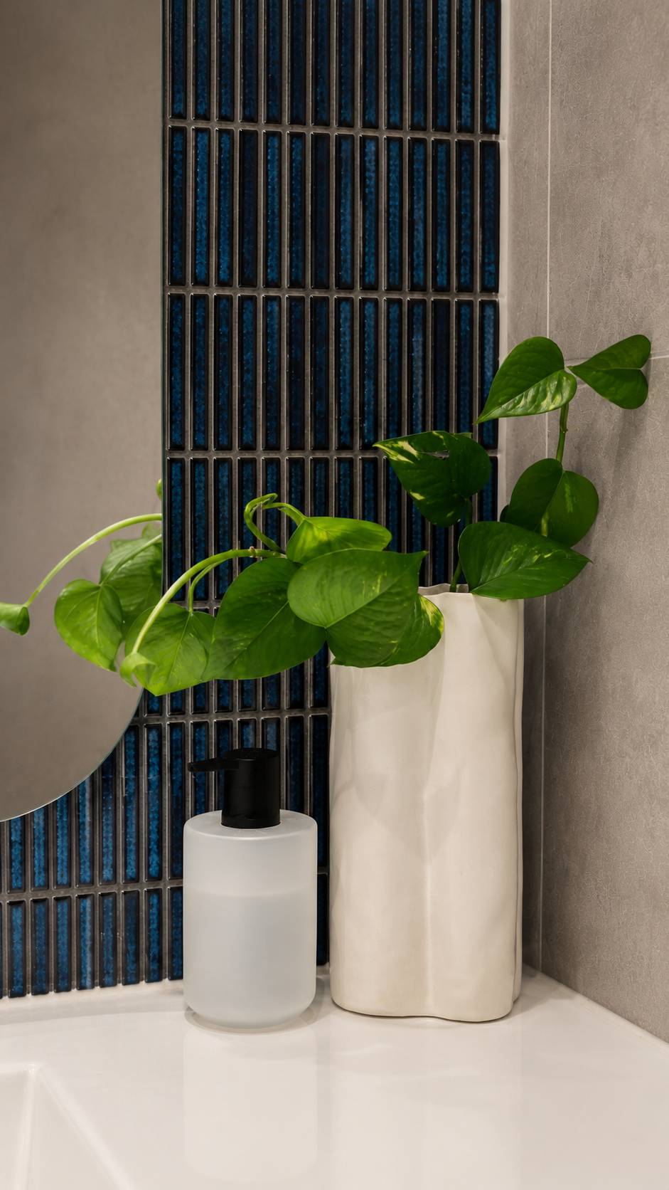 A tall white ceramic vase with green pothos plant leaves, and a white soap dispenser with black pump on a bathroom counter with a tiled wall background.