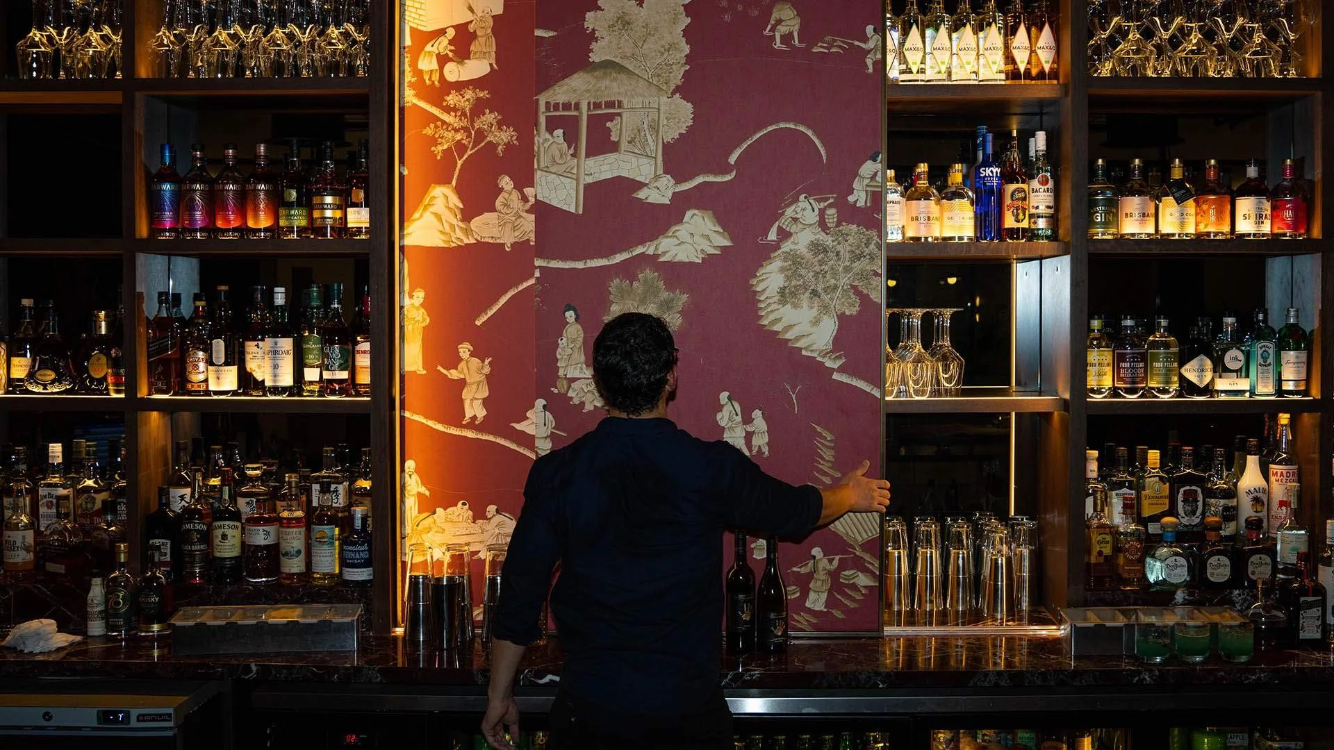 Bartender behind a bar preparing drinks in a dimly lit bar with shelves of liquor bottles and a decorative wall mural in the background.