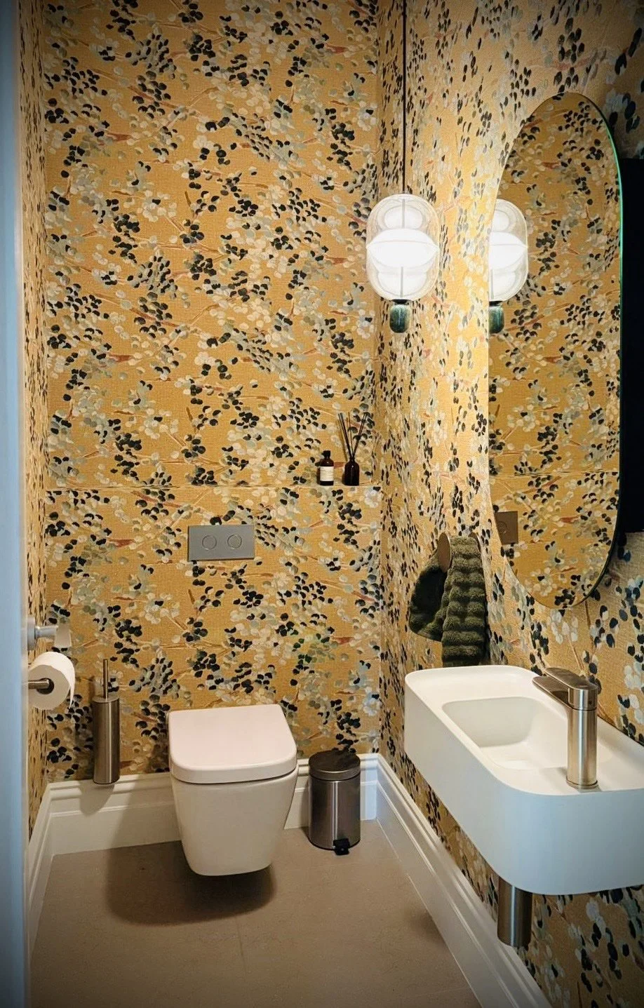 Small bathroom with patterned yellow wallpaper, a wall-mounted toilet with a toilet paper holder and waste bin, a narrow wall-mounted sink with a modern faucet, a large round mirror, and wall-mounted lights with white glass globes.