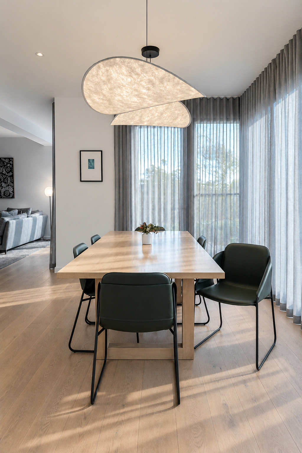 Modern dining room with a wooden table, six green chairs, large windows with sheer gray curtains, and a cloud-shaped ceiling light fixture.