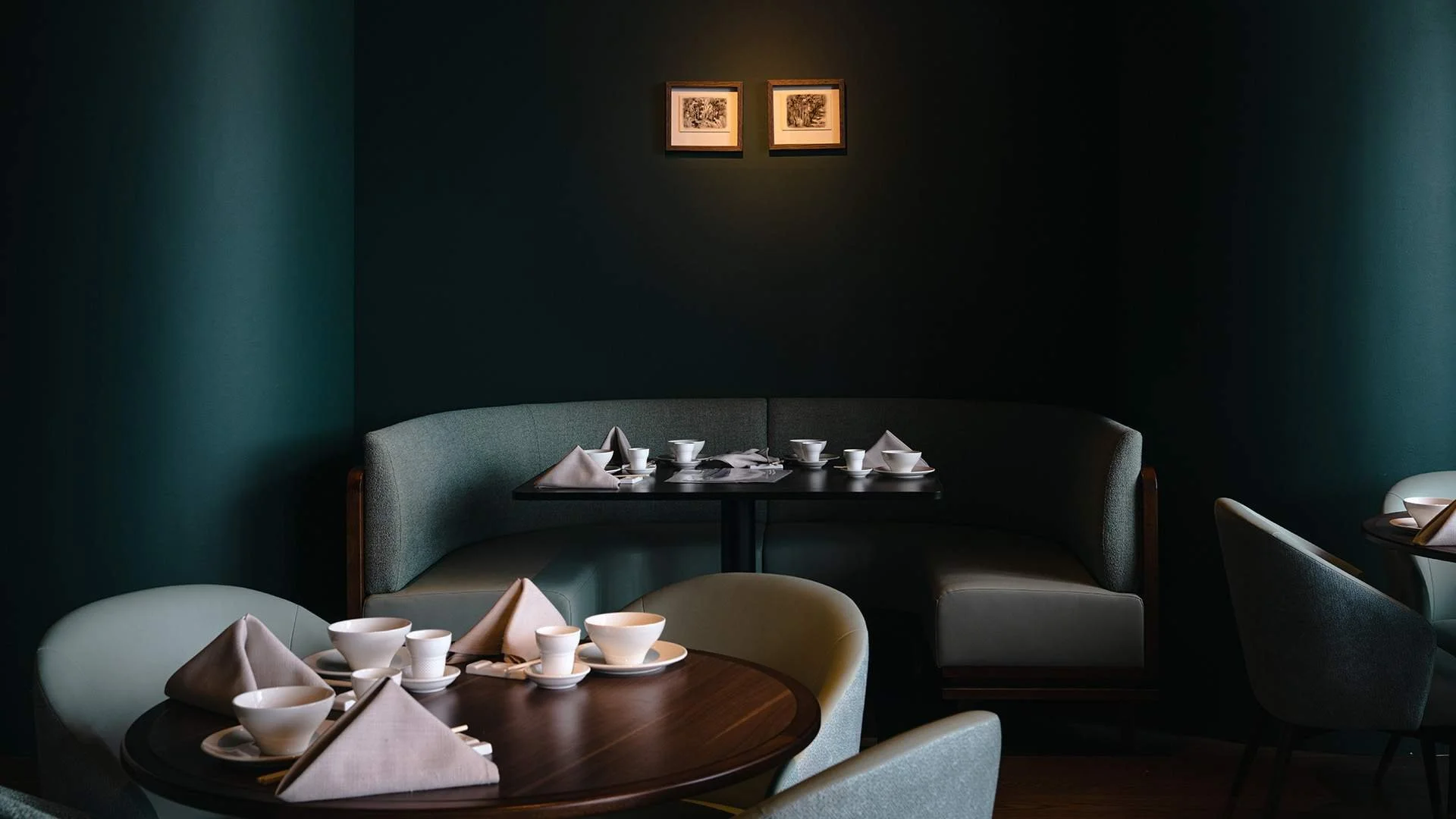Empty restaurant dining area with dark green walls, round tables set with white bowls, cups, napkins, and utensils, and framed artwork on the wall.