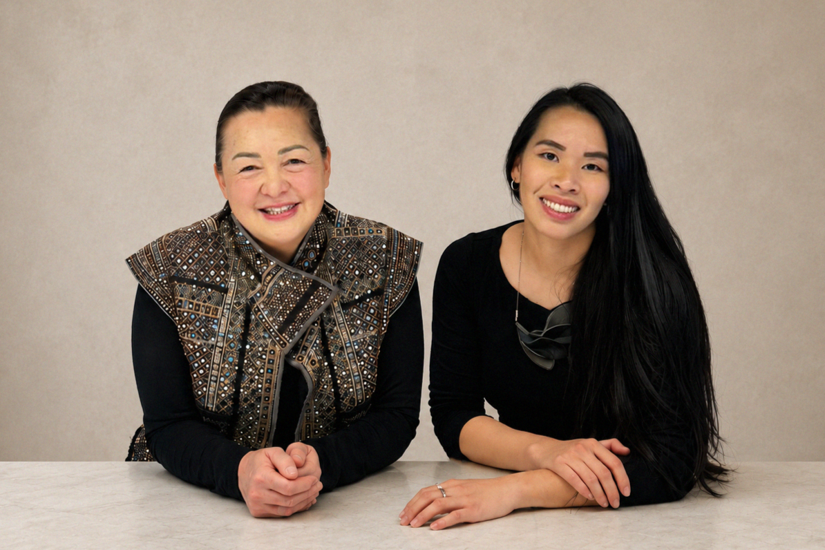 Two women sitting at a table, smiling at the camera. The woman on the left is older, wearing a patterned vest with a bow, and the woman on the right is younger, with long black hair and wearing a black top and jewelry.