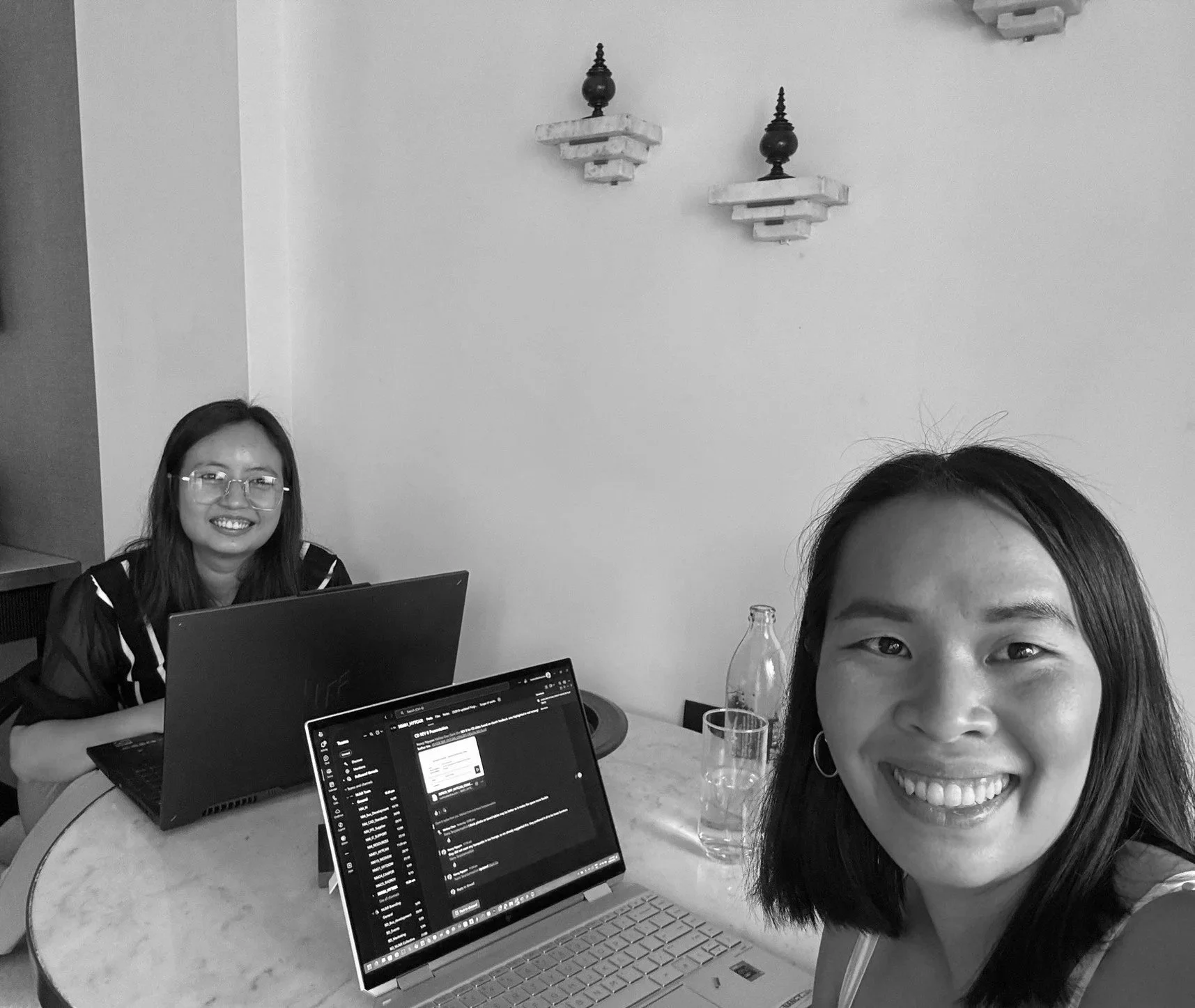 Two women smiling with laptops and drinks at a table, with one woman partially visible in the foreground and the other woman in the background.