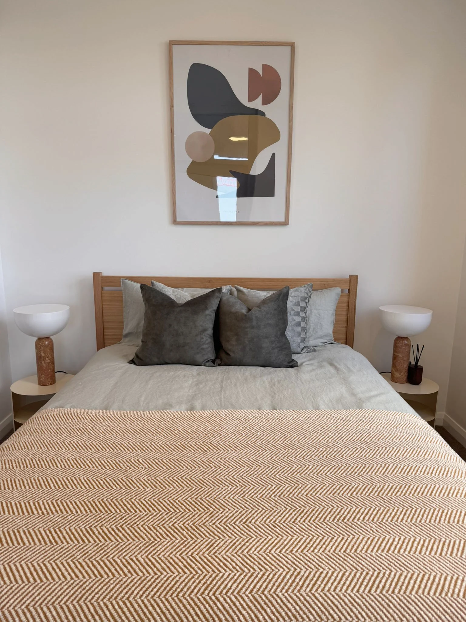 A cozy bedroom with a wooden bed frame, neutral bedding, and four pillows, two dark gray and two light. Two matching white bedside tables with granite bases and white lamps are on each side of the bed. A modern abstract art piece hangs above the bed 