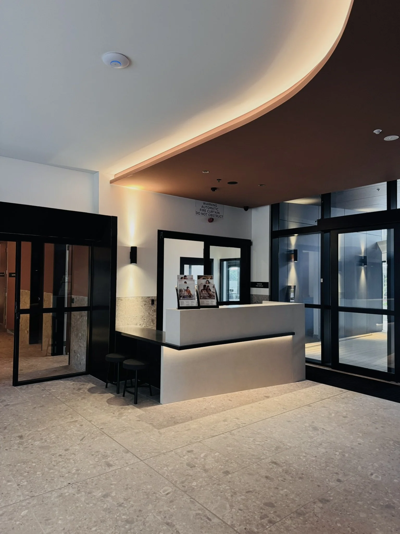 Modern hotel lobby with check-in counter, black stools, glass entrance doors, and subdued lighting.