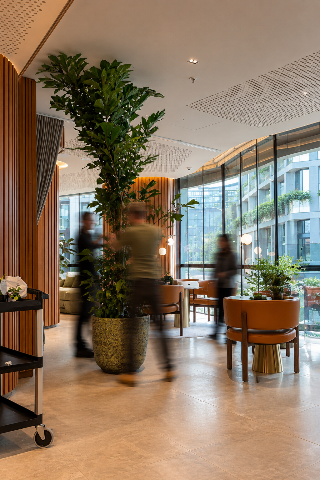 Modern cafe interior with large potted plant, wooden accents, brown chairs, large glass windows, and blurred people walking.