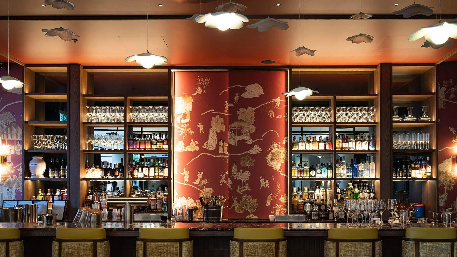 A bar with a wooden counter, bar stools, and backlit shelves filled with various bottles and glassware. Decorative wall art and ceiling lights shaped like clouds.