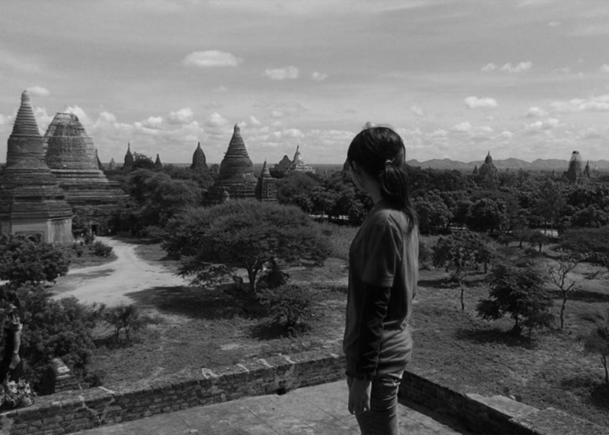 A person with long hair tied back, wearing a jacket, stands overlooking ancient temple ruins with pagoda-style structures under a partly cloudy sky.