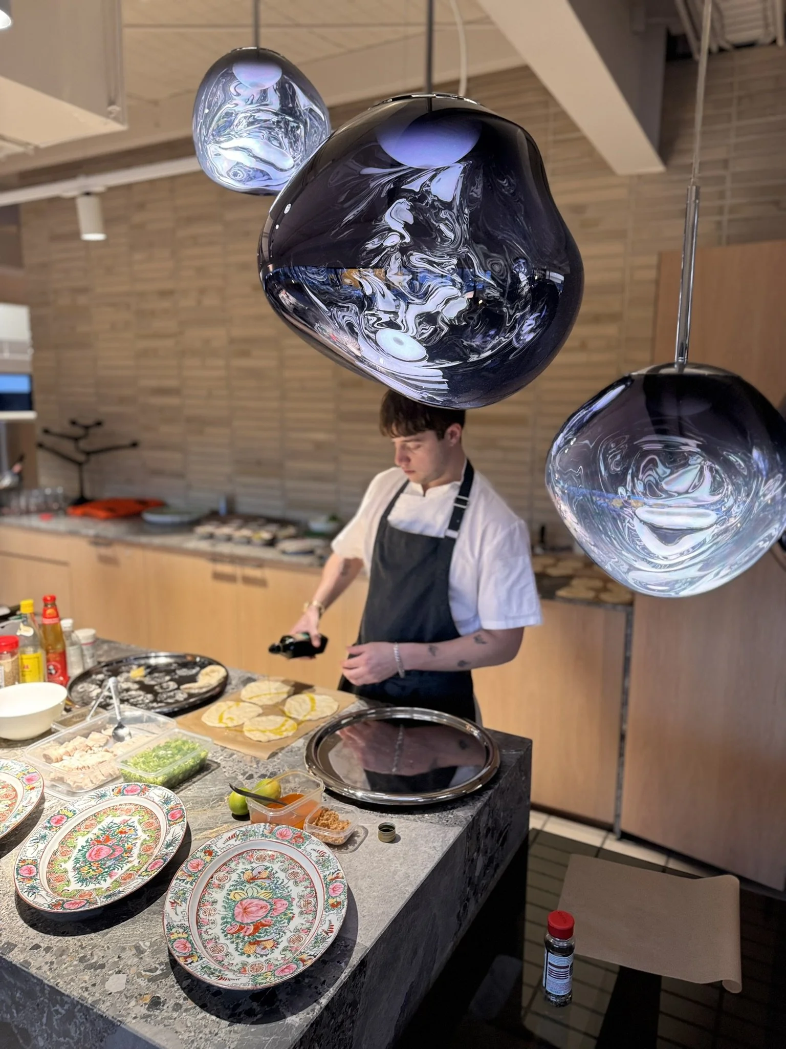Restaurant kitchen with chef preparing food, decorated with colorful patterned plates, vegetables, and sauces on the counter, and elegant hanging glass fixtures overhead.