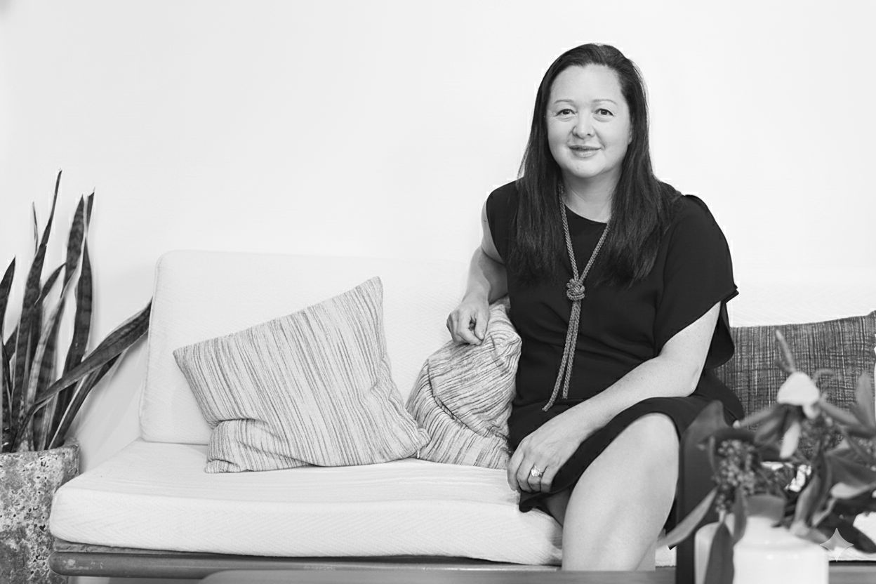 A woman sitting on a white sofa with striped pillows, smiling, with a potted plant on the left and a flower arrangement on the right.