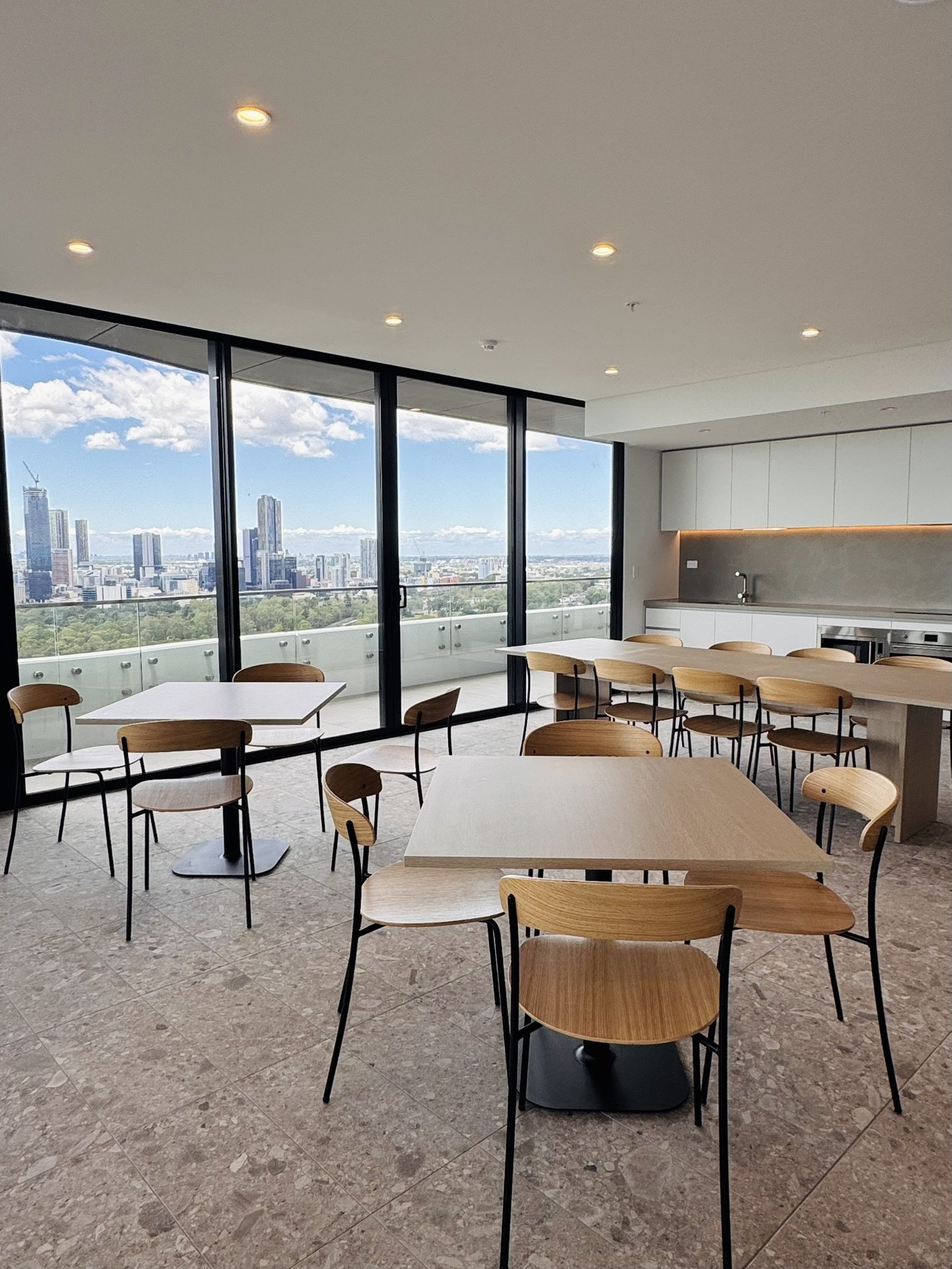 Modern dining area with wooden chairs and tables, large glass windows showing a city skyline and blue sky outside.