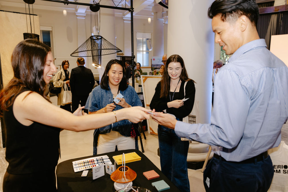 A group of four people at a professional event, with three women and one man, engaging around a table with promotional materials, in a well-lit room with other attendees in the background.