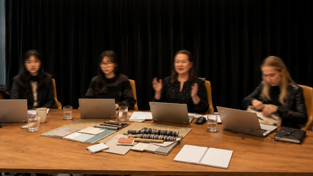 Four women sitting at a conference table with laptops, notebooks, fabric samples, and glasses of water, engaged in a discussion in a professional setting.