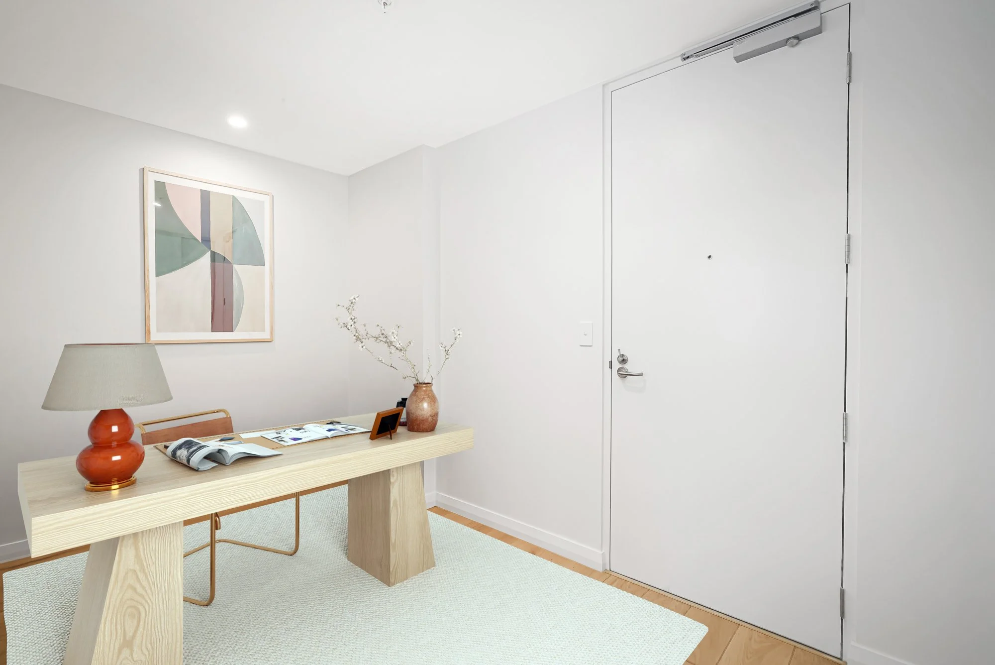 A minimalist entryway with a light wood table, orange table lamp, open magazine, framed artwork, glass vase with white branches, and a white door with a door closer, all in a clean white space.