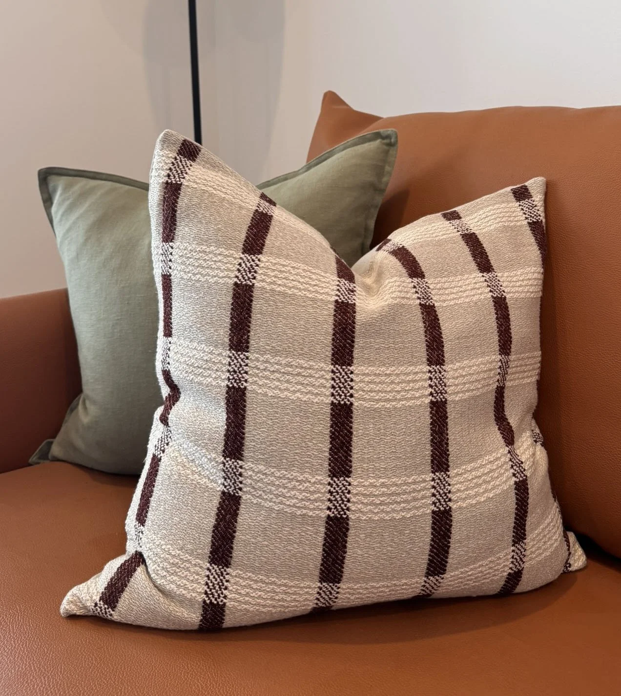 A brown checkered decorative pillow on a tan leather sofa, with two green throw pillows in the background.