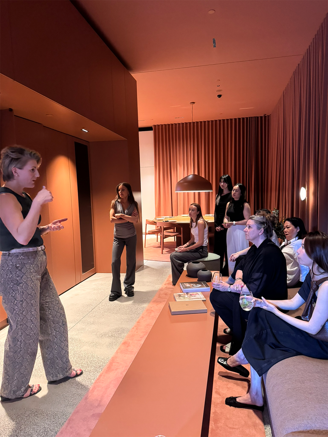 A group of women are seated and standing in a modern, warmly lit room, listening attentively to a woman who appears to be speaking or giving a presentation. The room features wooden paneling, a pinkish-brown decorative wall, and a table with books and other items.