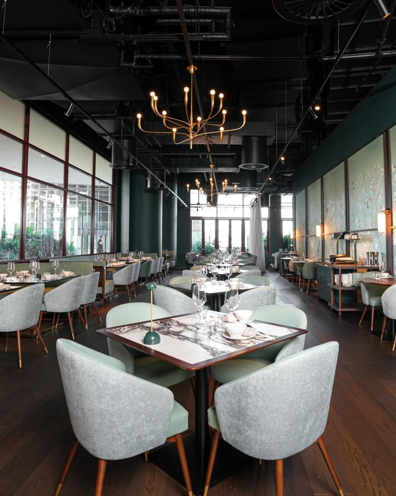 Modern upscale restaurant interior with large windows, elegant tables set with glassware and bowls, comfy gray chairs, gold chandeliers, and ambient lighting.
