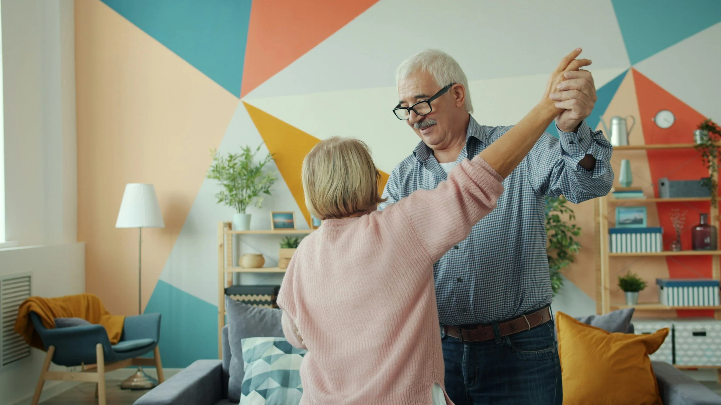 An elderly man and woman dancing together in a cozy living room with colorful geometric wall art, plants, and decorative shelves.