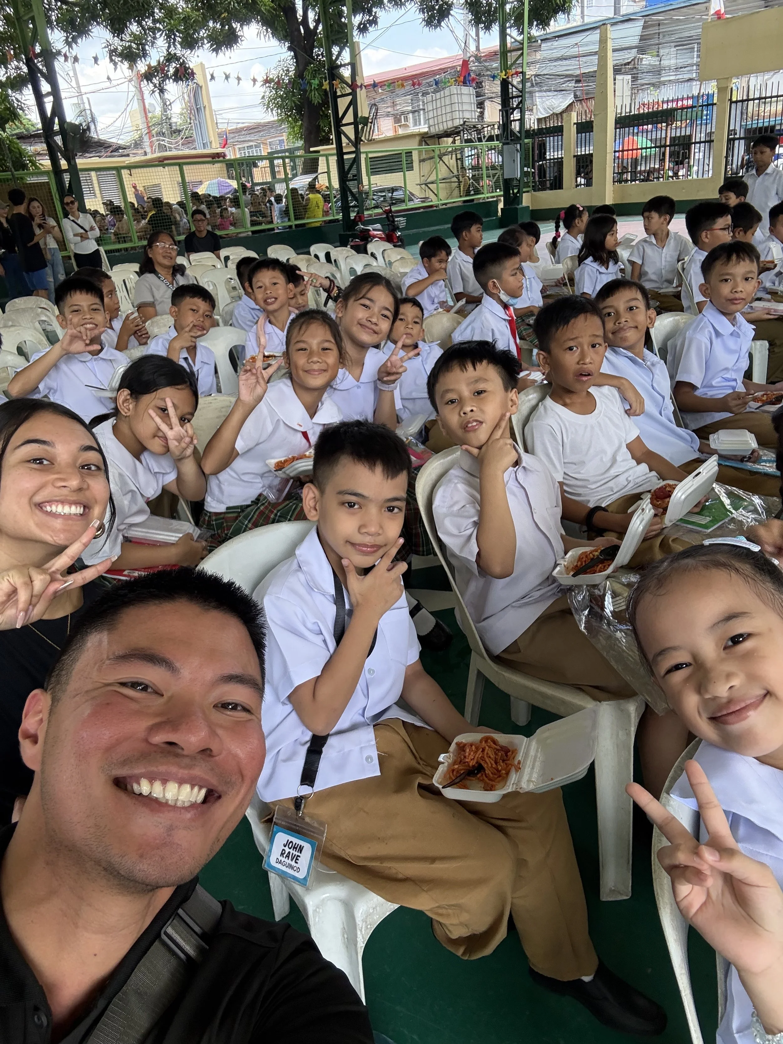 A group of school children in uniforms sitting at tables, eating lunch, with some making peace signs and smiling for a photo taken by an adult male in the foreground.