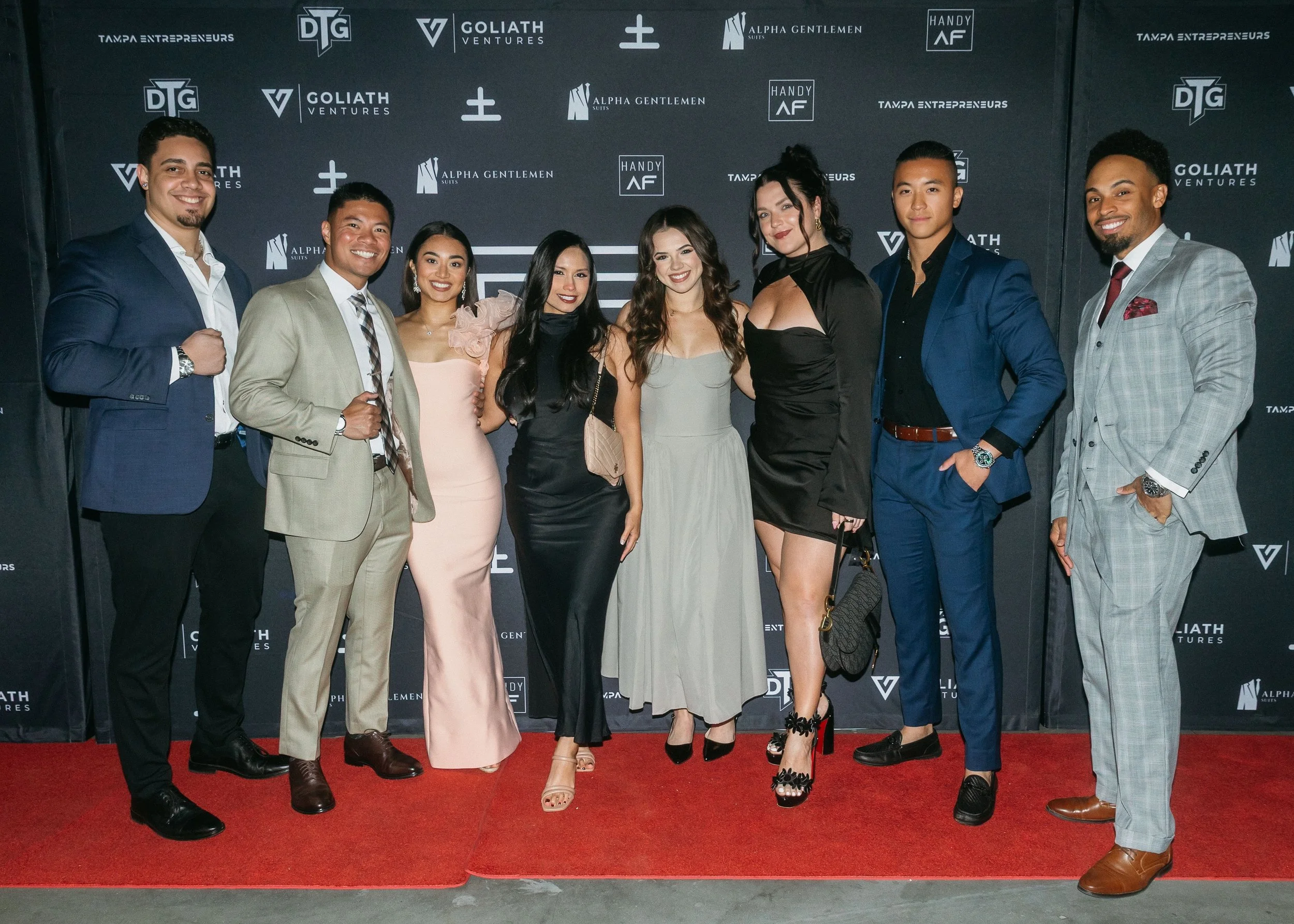 Group of nine diverse young adults dressed in formal attire, posing on a red carpet at an event with a branded backdrop featuring logos like Tampa Entrepreneurs, Goliath Ventures, Alpha Gentlemen, and Handy AF.