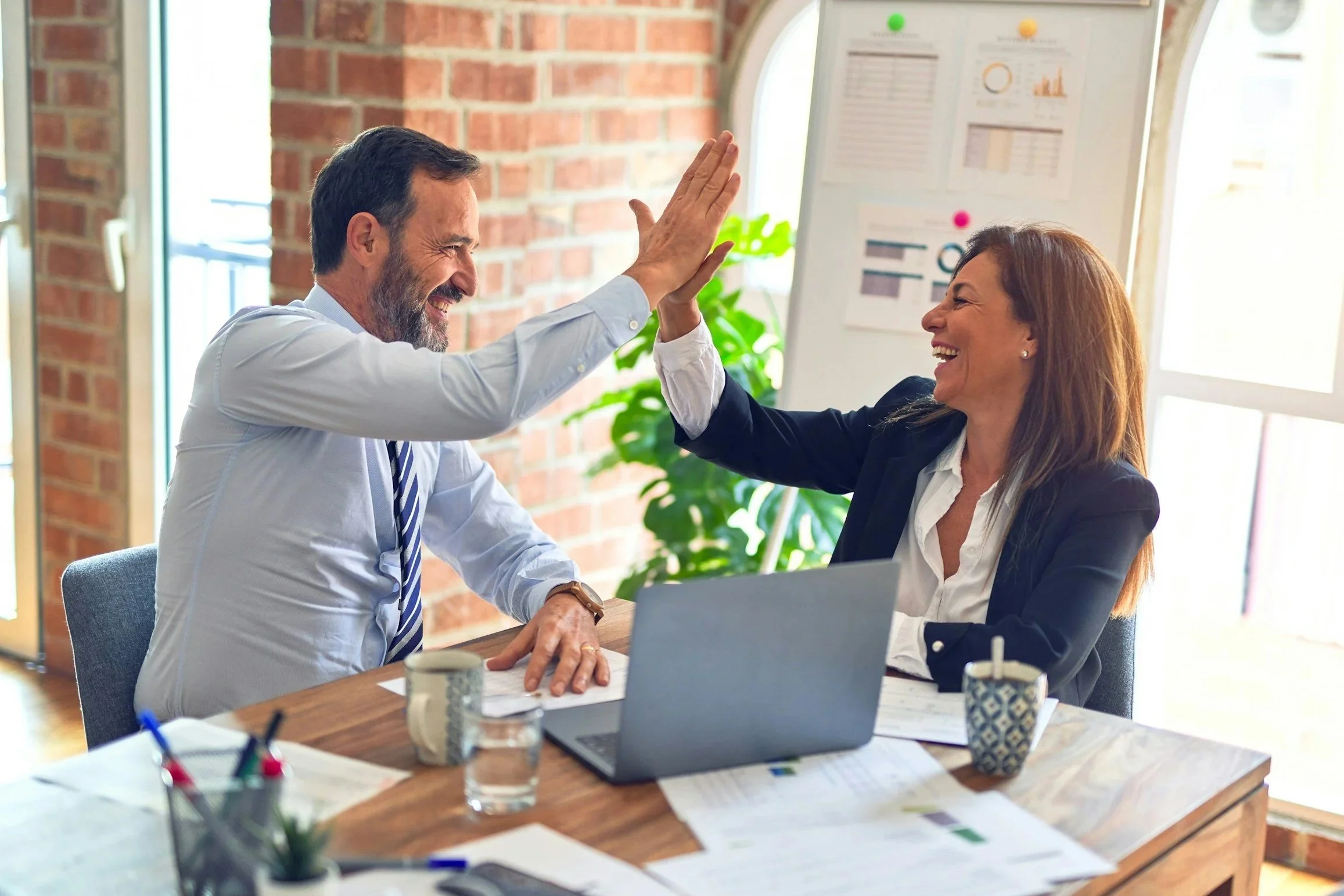 A man and woman sharing a high five in a bright, casual office setting, sitting at a wooden table with a laptop, papers, and coffee mugs, with a brick wall and window in the background.