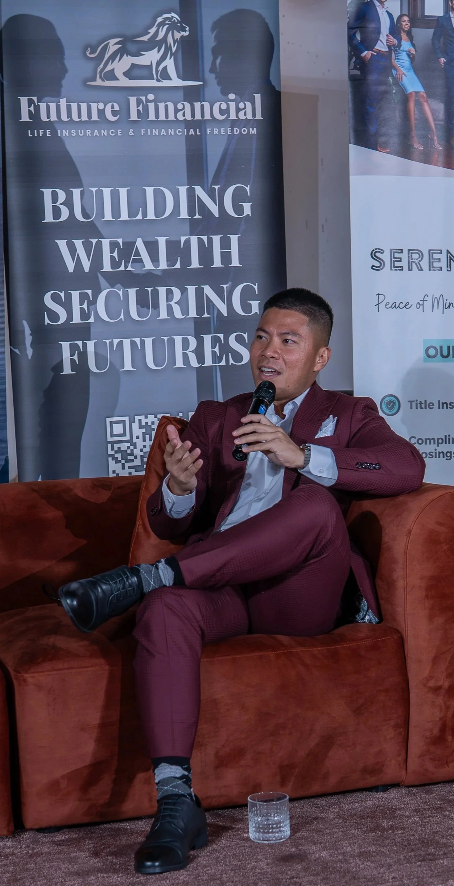 A man in a maroon suit speaking into a microphone while sitting on a orange sofa, with a financial seminar backdrop that has a lion logo and the words: 'Future Financial - Building Wealth Securing Futures'.