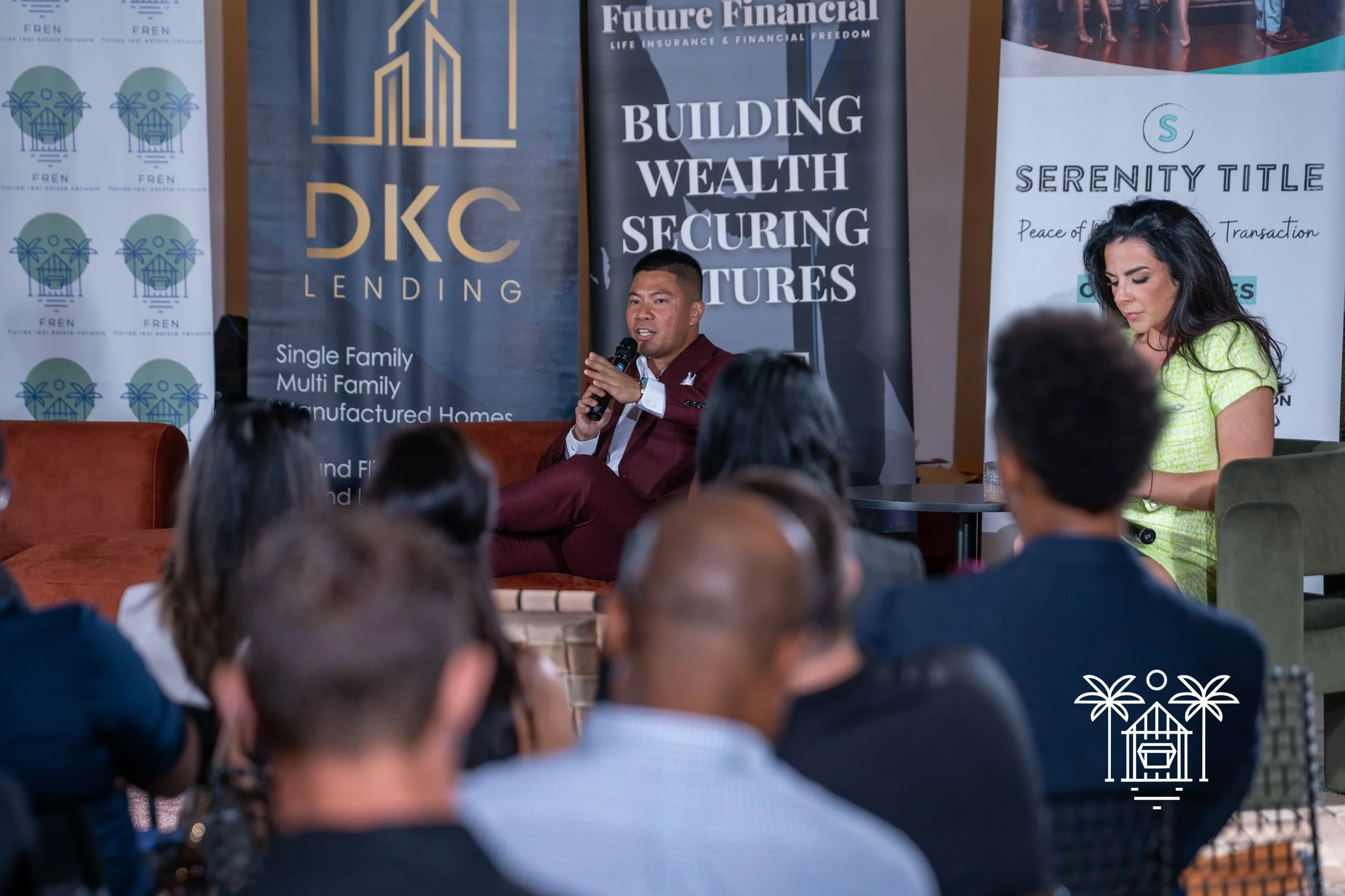 A man in a maroon suit and a woman in a yellow dress are speaking at a financial seminar. The audience is seated facing them, and there are various promotional banners in the background related to financial services and real estate.