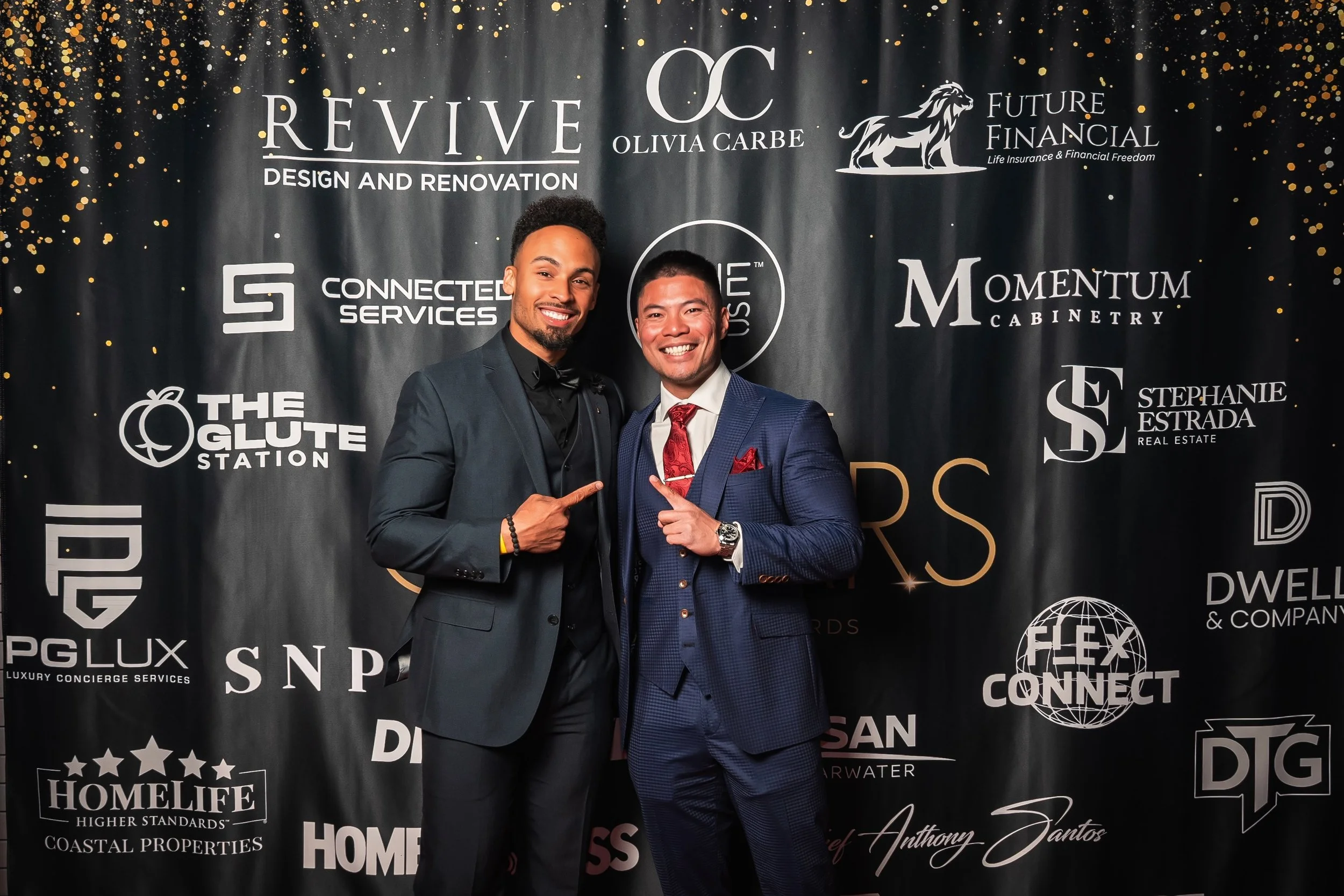 Two men in formal suits smiling and pointing at each other in front of a black backdrop with gold splatter and company logos, including 'Revive Design and Renovation,' 'Olivia Carbe,' and 'Future Financial.'