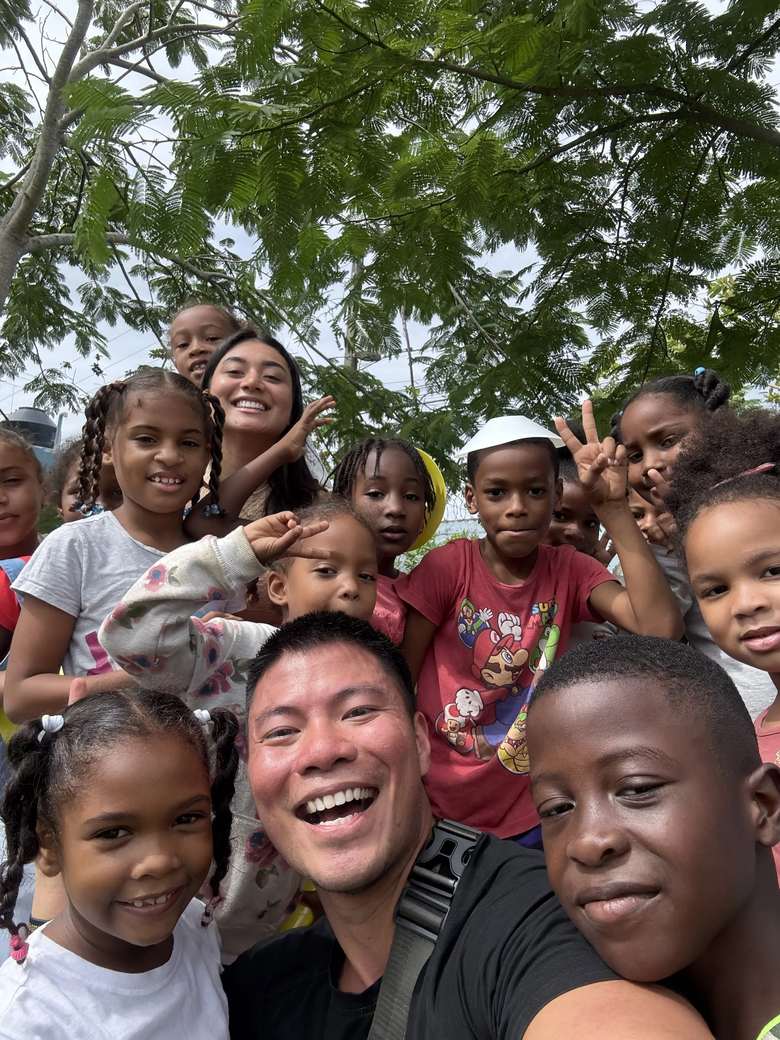 Smiling group of children and a man taking a selfie outdoors under a large green tree.