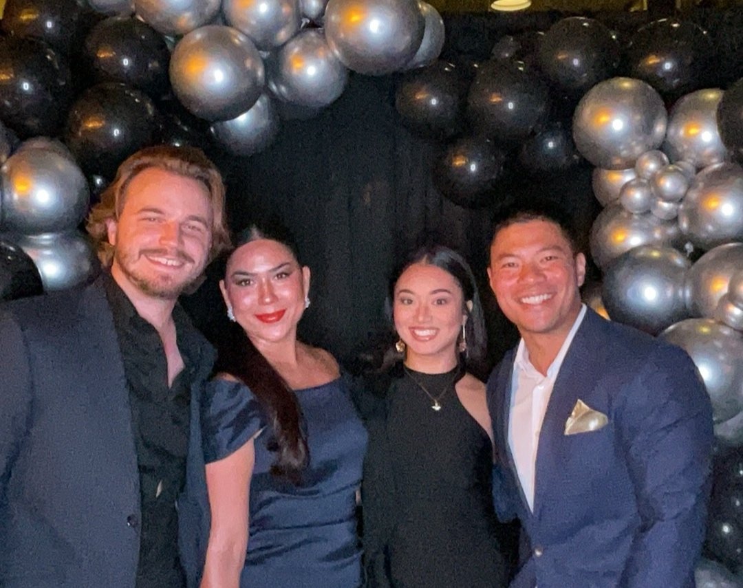 Four people dressed in formal attire smiling at a party with black and silver balloons in the background.