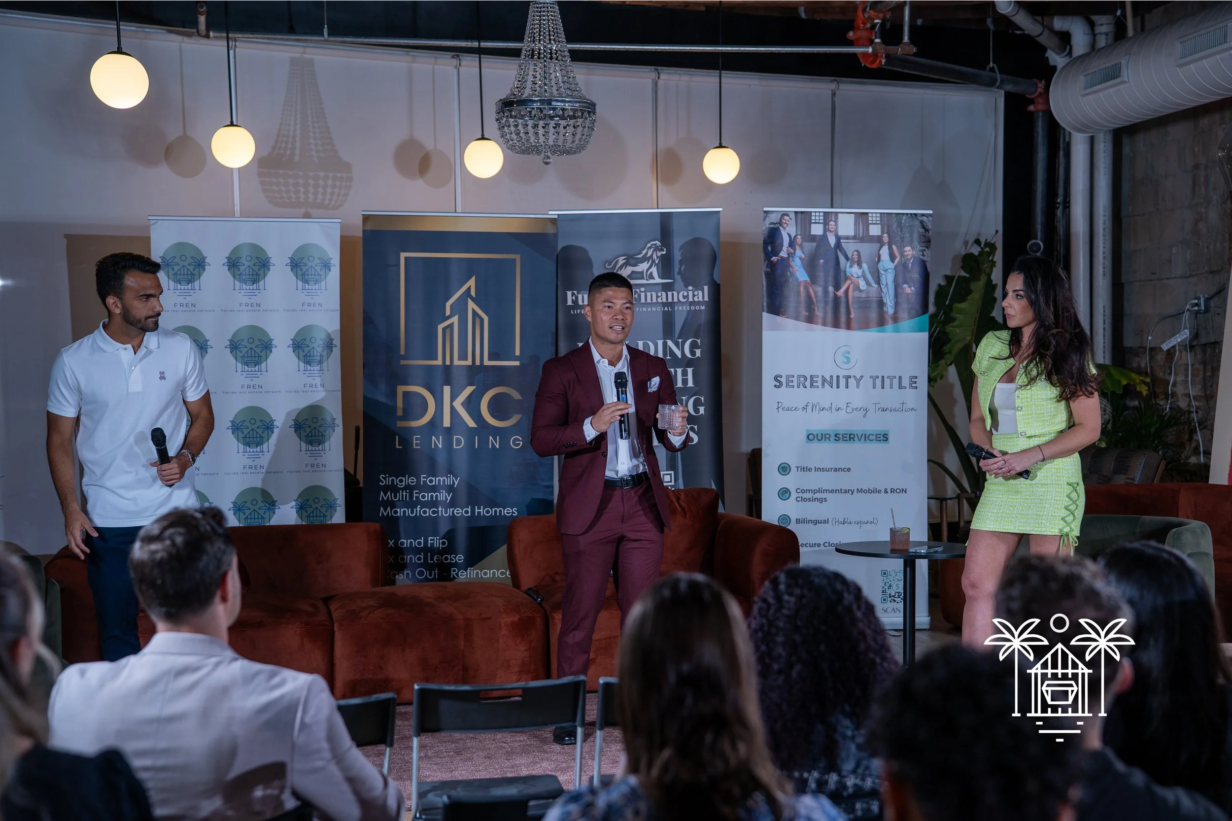 Three people on a stage speaking to an audience during a business event. The man in the center is wearing a burgundy suit and holding a microphone. A woman on the right in a light green dress and a man on the left in a white shirt also hold microphones. Behind them are banners with logos and text, and there are audience members seated watching.