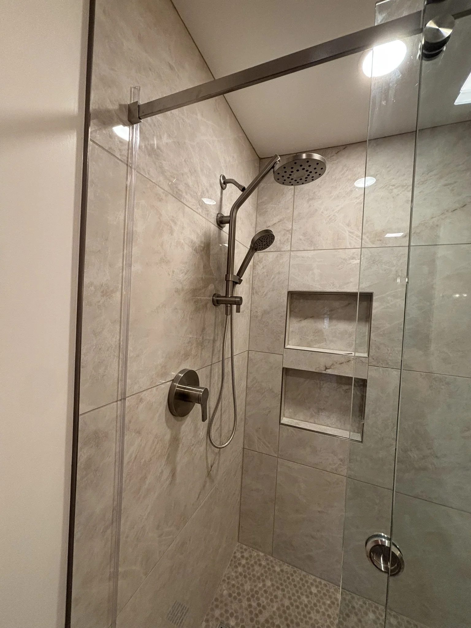 A modern walk-in shower with beige marble walls, a round overhead shower head, a handheld shower head, a built-in tiled niche with two shelves, a glass barn door, a small drain on the beige tiled floor, and a shiny chrome fixtures.