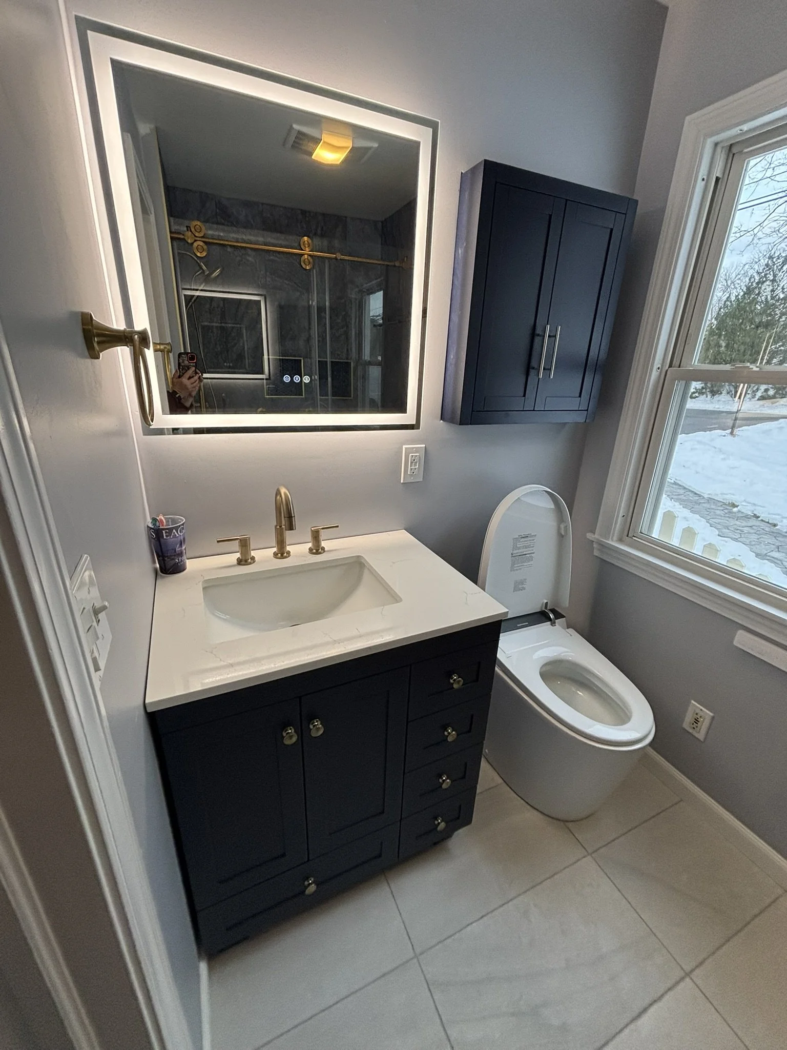 Bathroom with navy blue vanity, marble countertop, and gold fixtures, mirror with backlighting, toilet, and window showing a snowy outdoor scene.