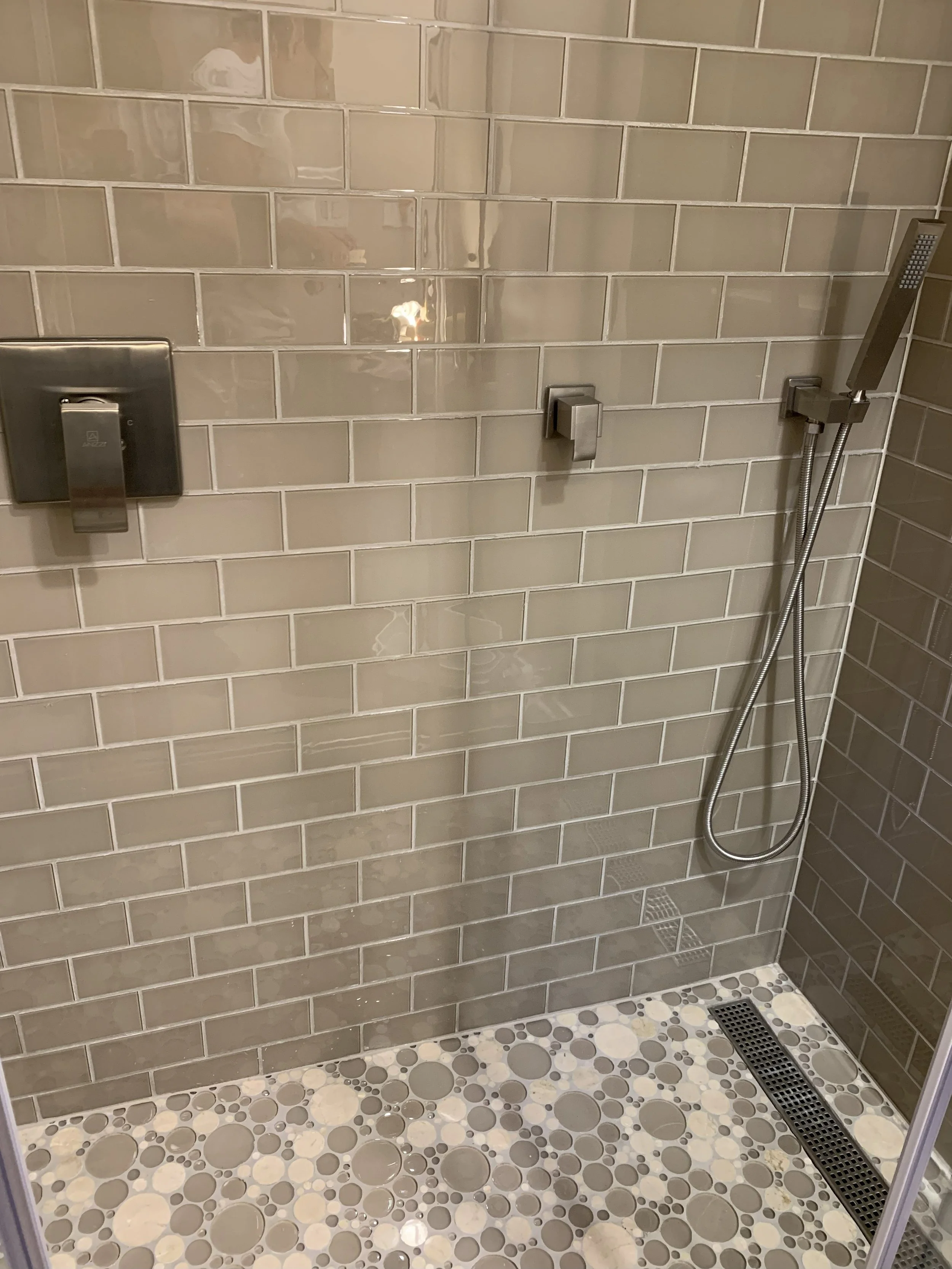 A modern shower area with beige subway tiles on the wall, a handheld shower head attached to a flexible hose, a square control valve, a wall-mounted switch, and a floor with multicolored bubble tiles and a linear trench drain.