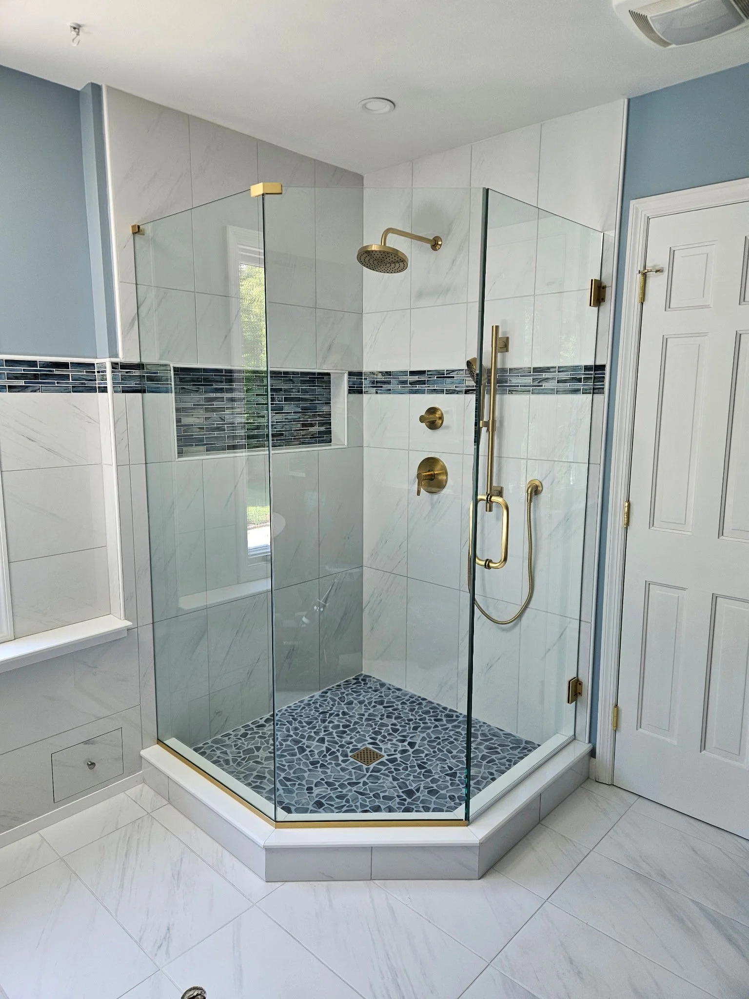 Modern glass-enclosed shower with brass fixtures, white marble tiles, and a mosaic glass tile floor in a bathroom with matching white marble and a white door.