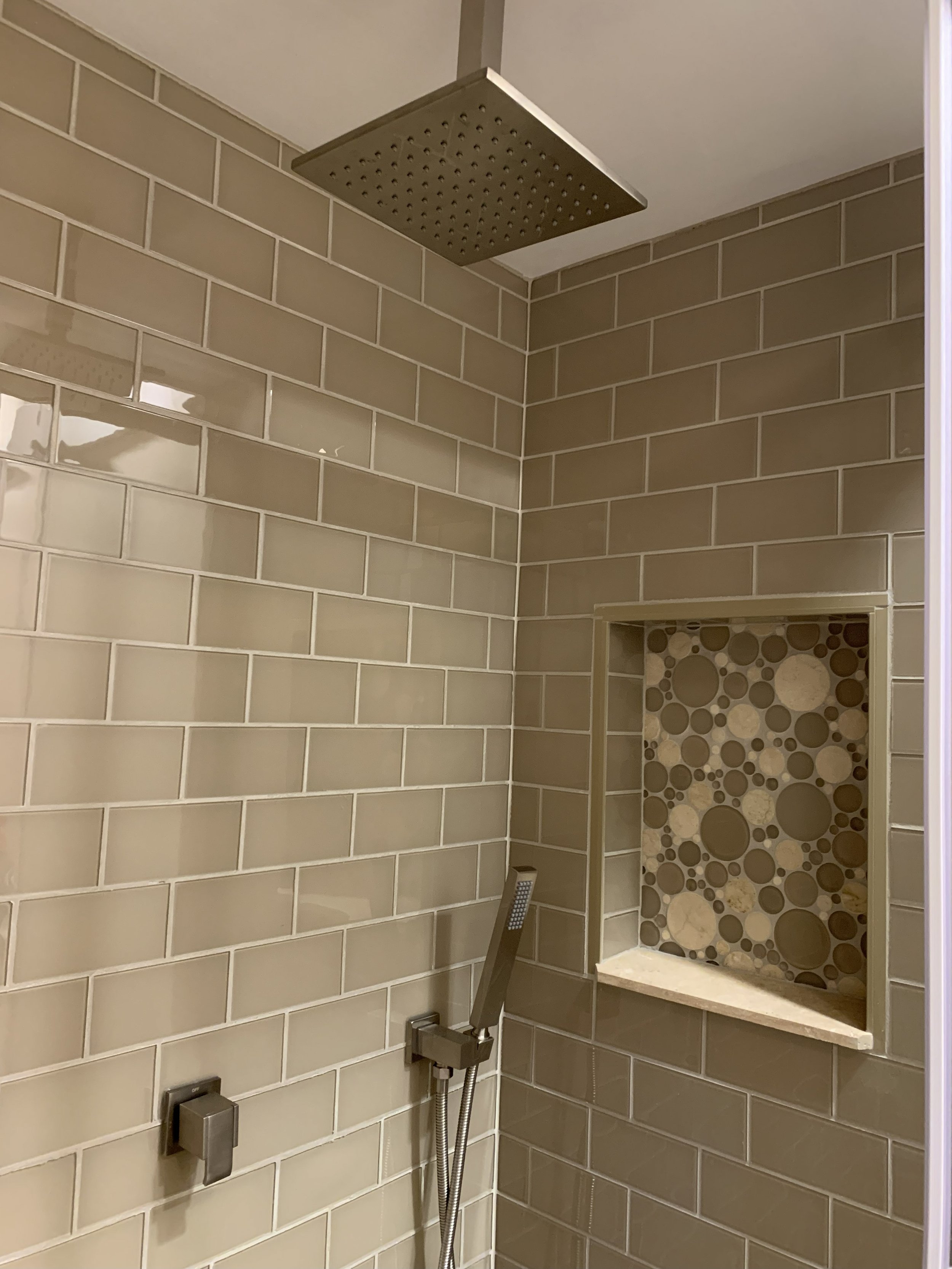 A shower area with beige subway tile walls, a rain showerhead, a handheld shower, and a built-in niche with round stone mosaic tiles.