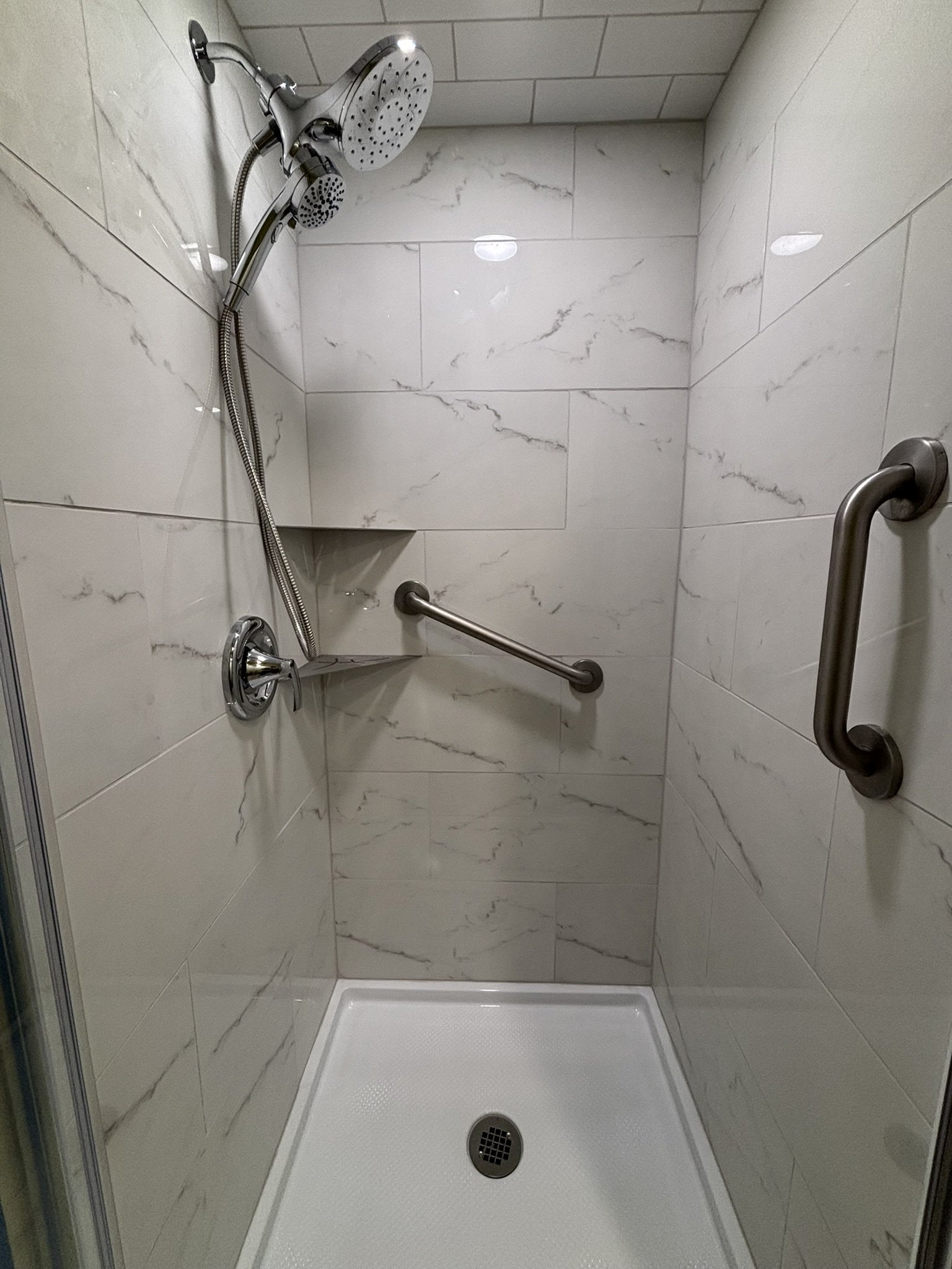A walk-in shower with white marble-patterned tiles on the walls, a handheld showerhead, a fixed showerhead, a grab bar, a small built-in corner shelf, and a textured shower floor with a drain in the center.