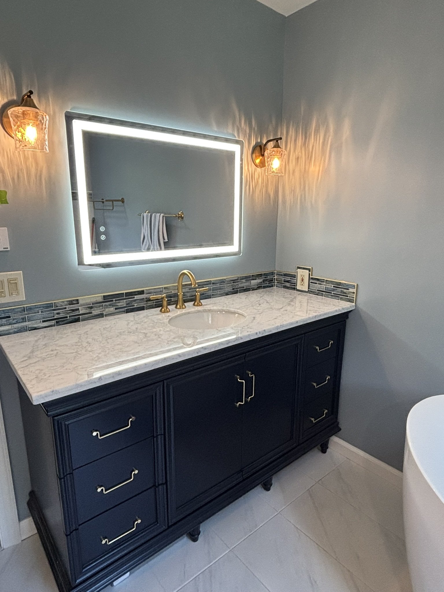 A bathroom vanity with a marble countertop, dark navy blue cabinets, gold hardware, a rectangular mirror with LED lighting, and a towel hanging on a gold towel bar. The wall is painted light blue and has two wall-mounted light fixtures with glass shades.
