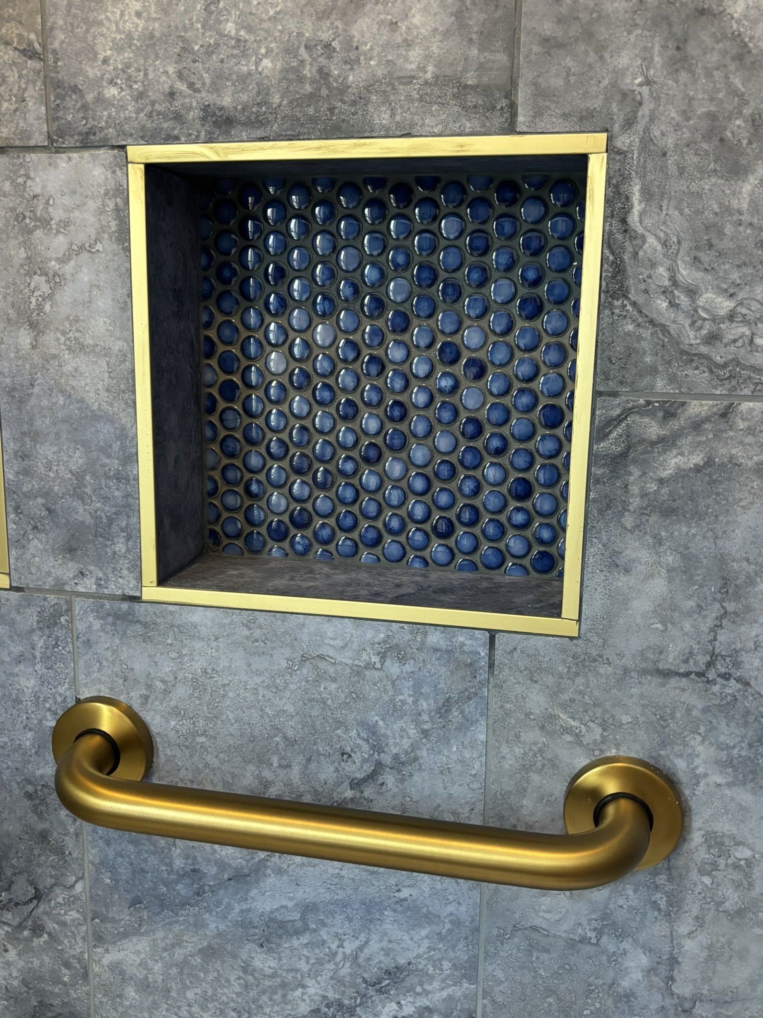 A built-in shower niche with aluminum trim and mosaic tile, surrounded by gray tiles and brushed-gold aluminum trim, with a gold-colored grab bar on the bottom.