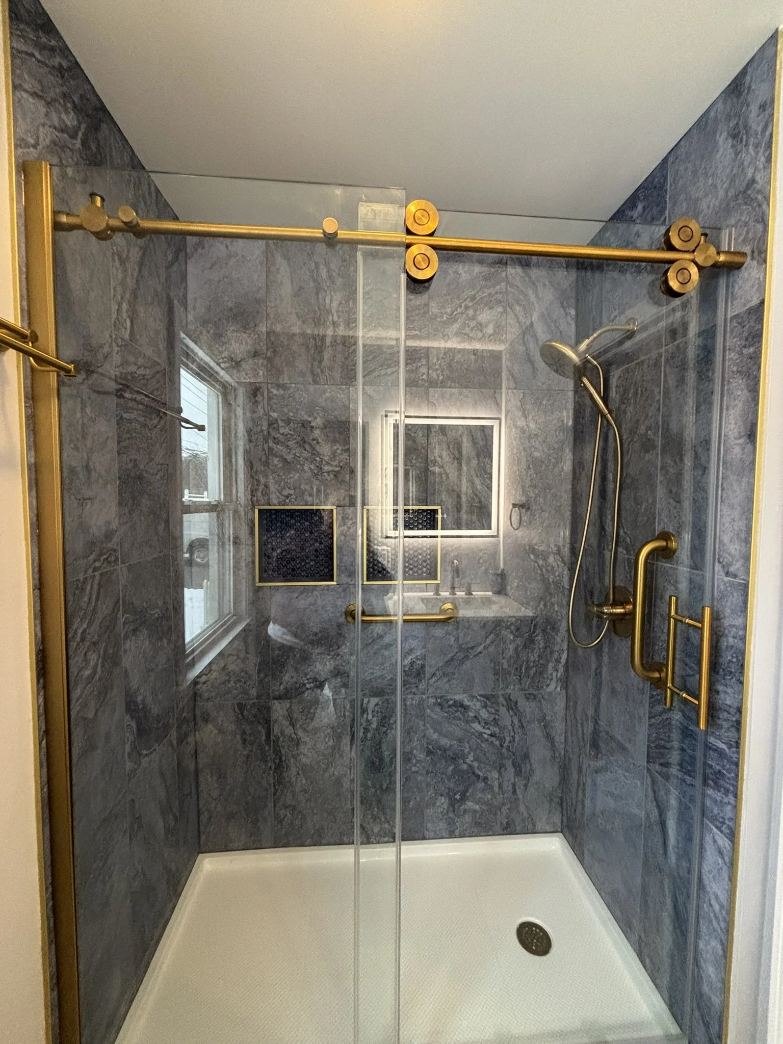 A modern shower with dark gray marble tiles, a glass door, brushed gold fixtures, a ceiling showerhead, and a window with niches.