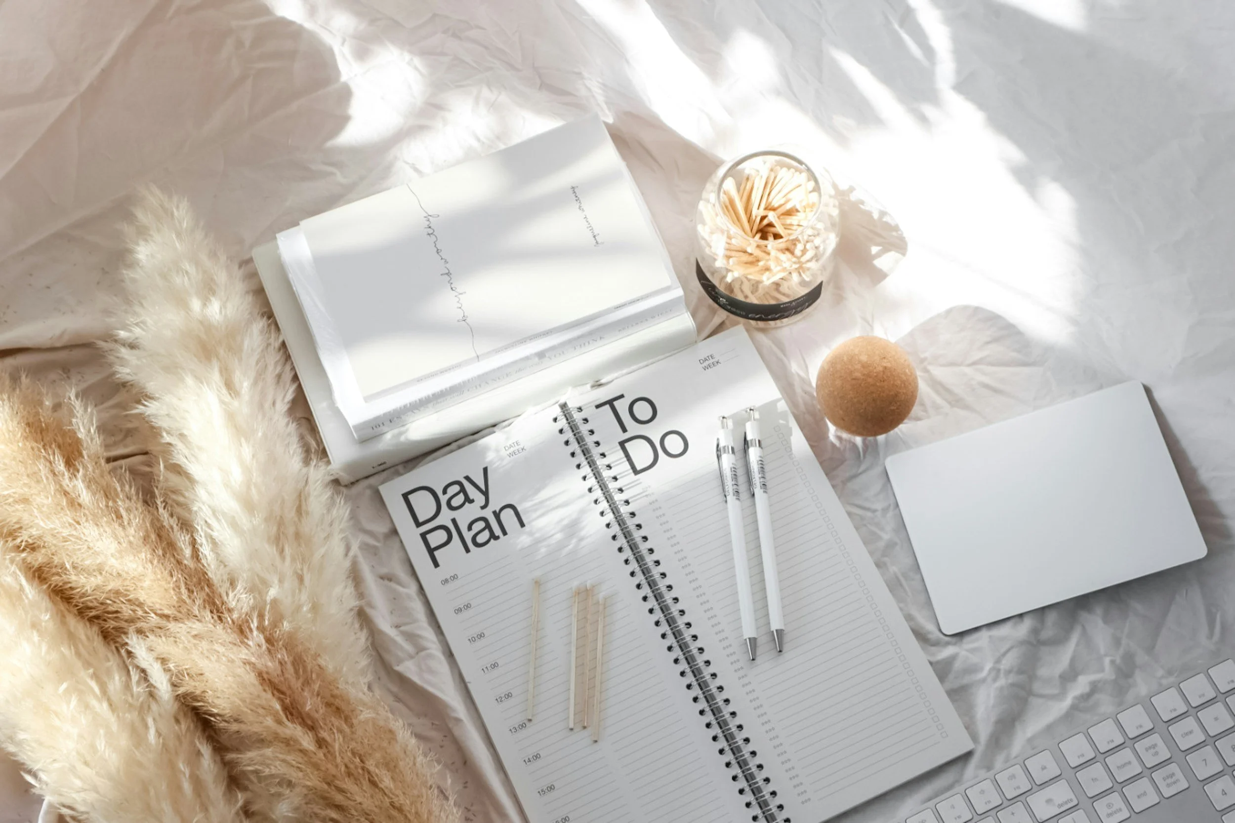 A flat lay of a workspace with a notebook, planner, pens, and a jar of toothpicks on crumpled white paper, illuminated by natural light, with a fluffy beige blanket on the side.