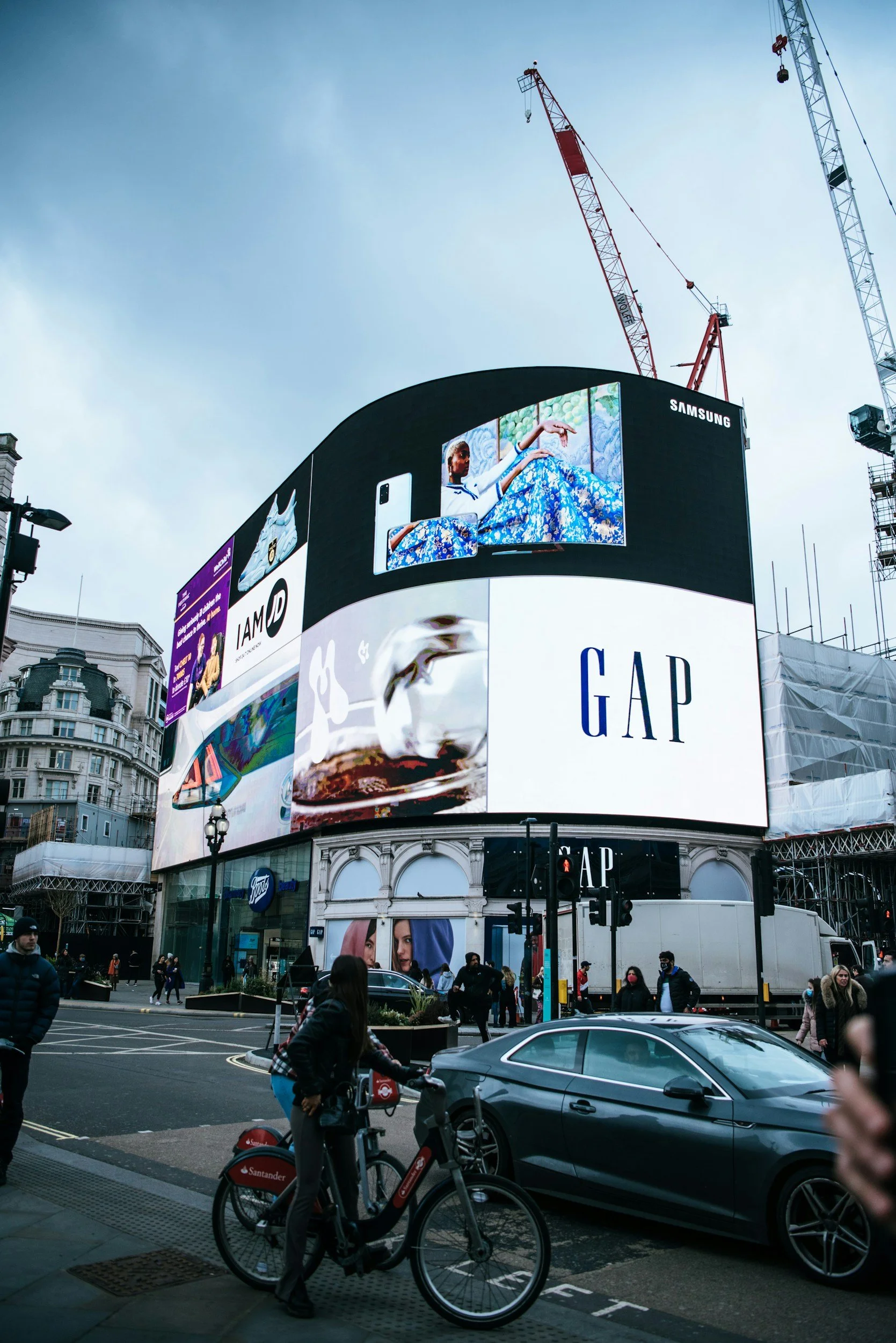 City street scene with digital billboards, pedestrians, a cyclist, and several buildings in the background.