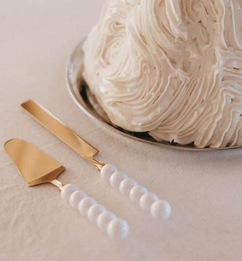 A silver bowl filled with a textured, creamy white cake with frosting swirls, alongside two gold-tined cake servers with white beaded handles on a beige surface.