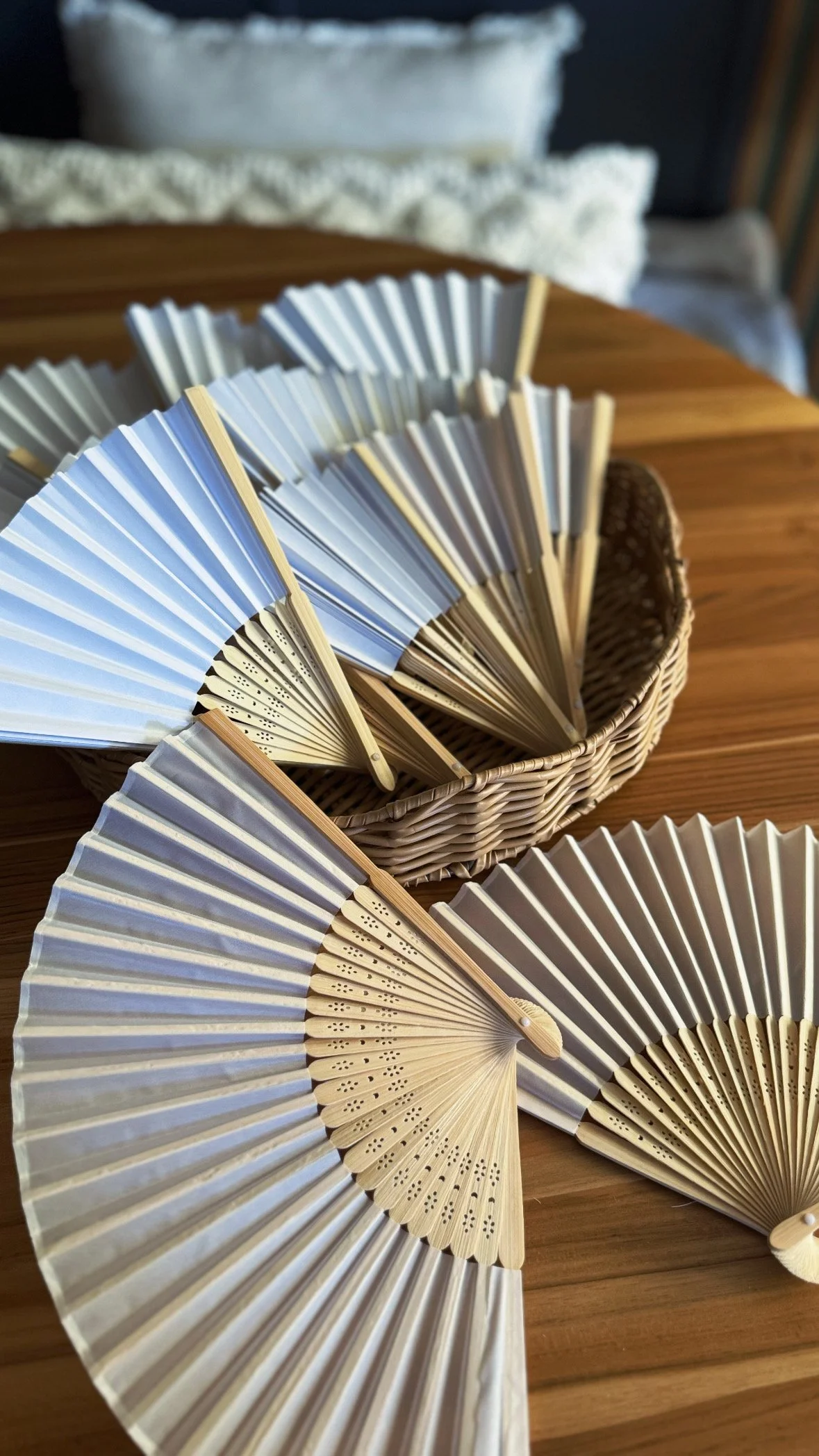 Multiple hand-held folding fans with wooden ribs and fabric or paper surfaces, some open and some closed, arranged in a basket and on a wooden surface.