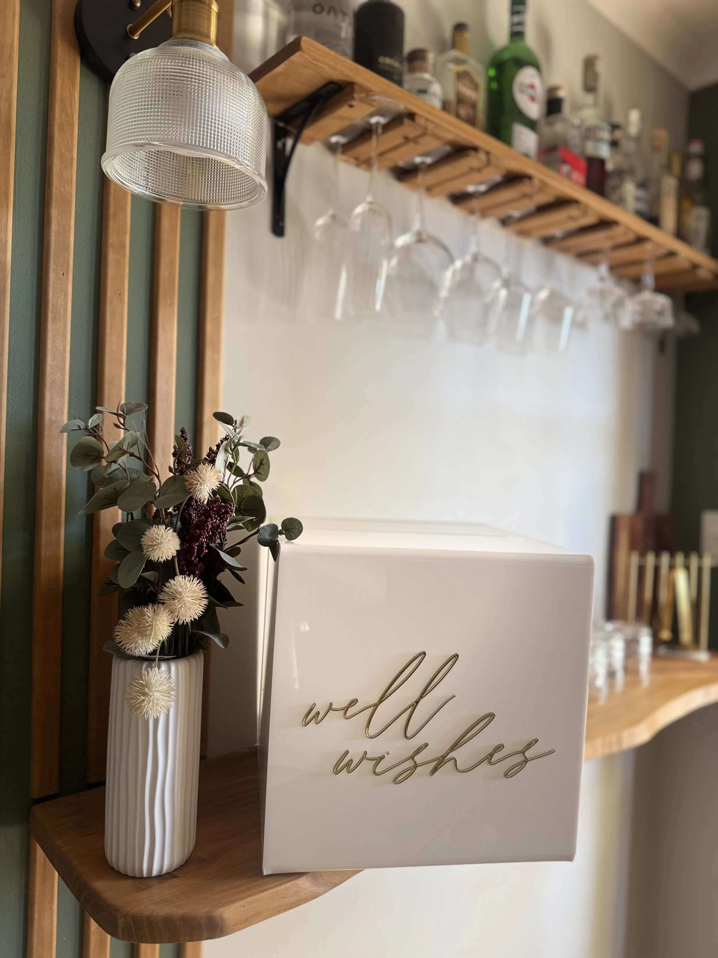 A white box with gold cursive writing that says 'well wishes' on a wooden shelf beside a white vase with dried flowers, with a wooden rack and hanging wine glasses in the background.